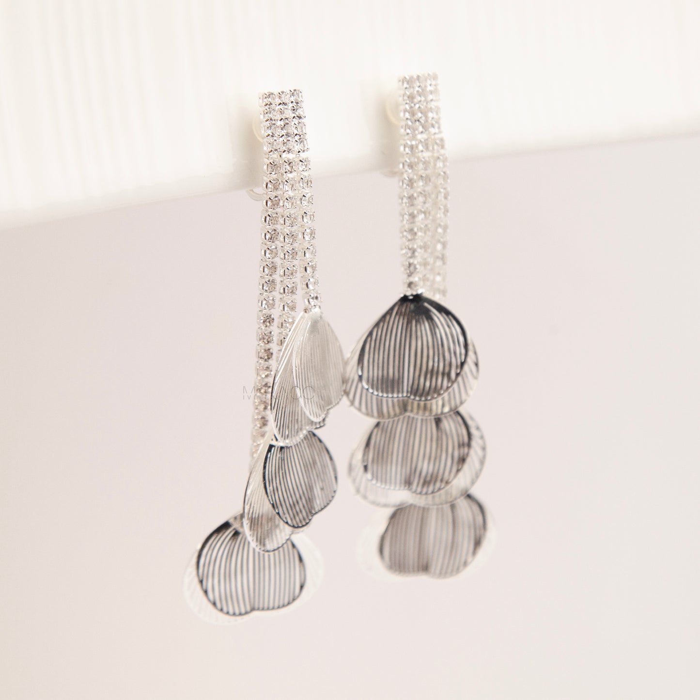 A pair of elegant silver dangle clip-on earrings, featuring three rows of sparkling rhinestones cascading into textured, hollow metallic leaf elements hanging in tiers.