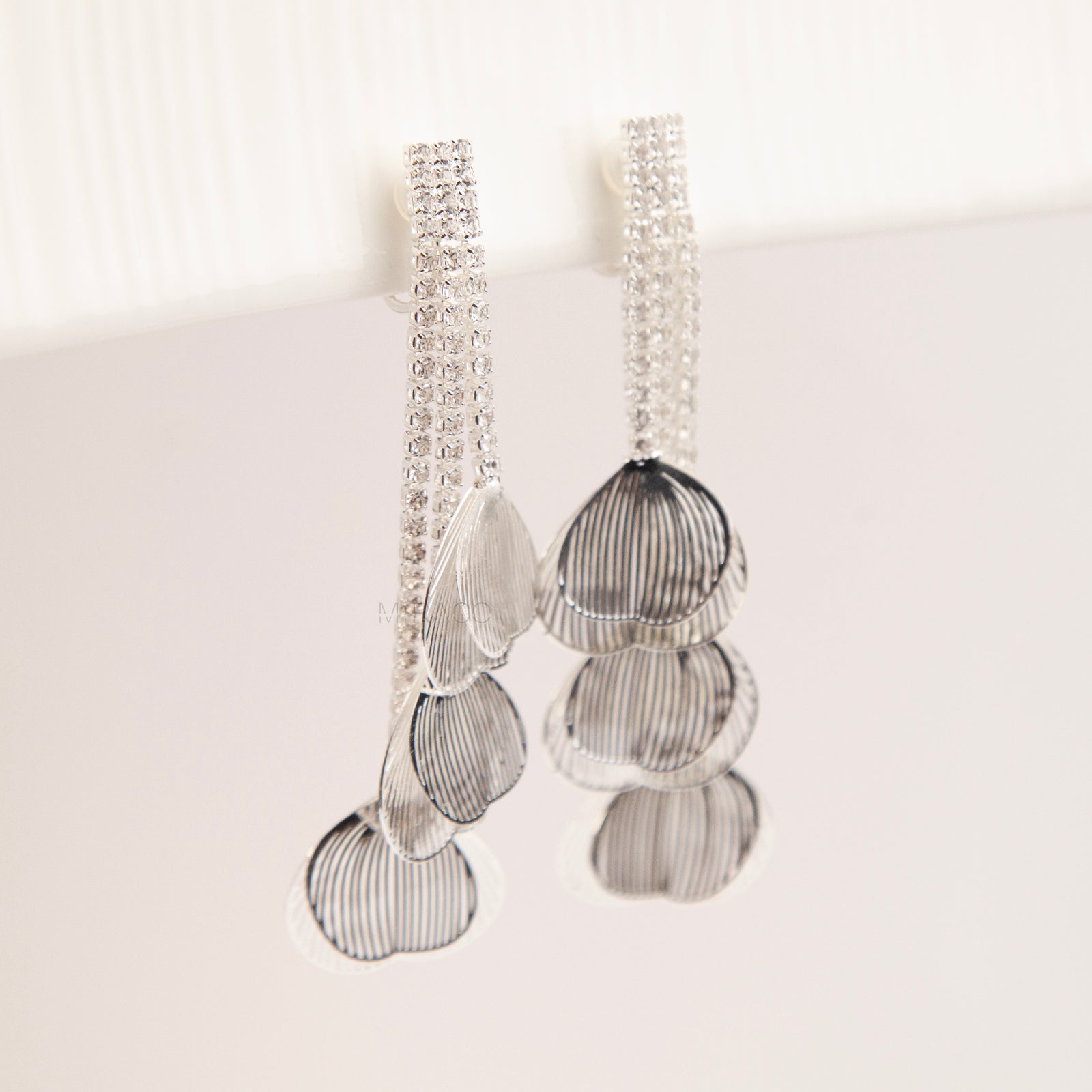 A pair of elegant silver dangle clip-on earrings, featuring three rows of sparkling rhinestones cascading into textured, hollow metallic leaf elements hanging in tiers.