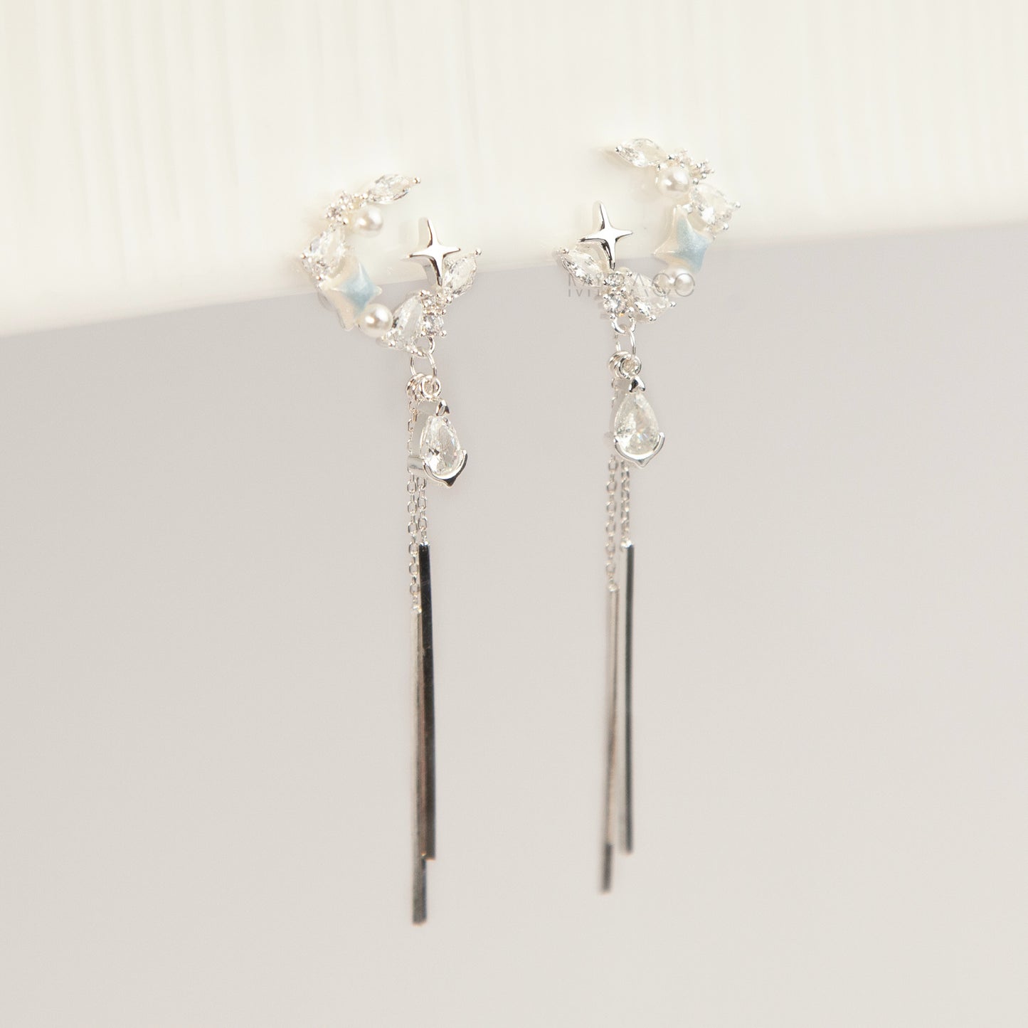 A pair of elegant silver celestial dangle clip-on earrings, featuring a crescent moon and star design accented with marquise crystals and tiny pearls, cascading into a teardrop gemstone and sleek, vertical metallic bar tassels on comfortable coil backs.