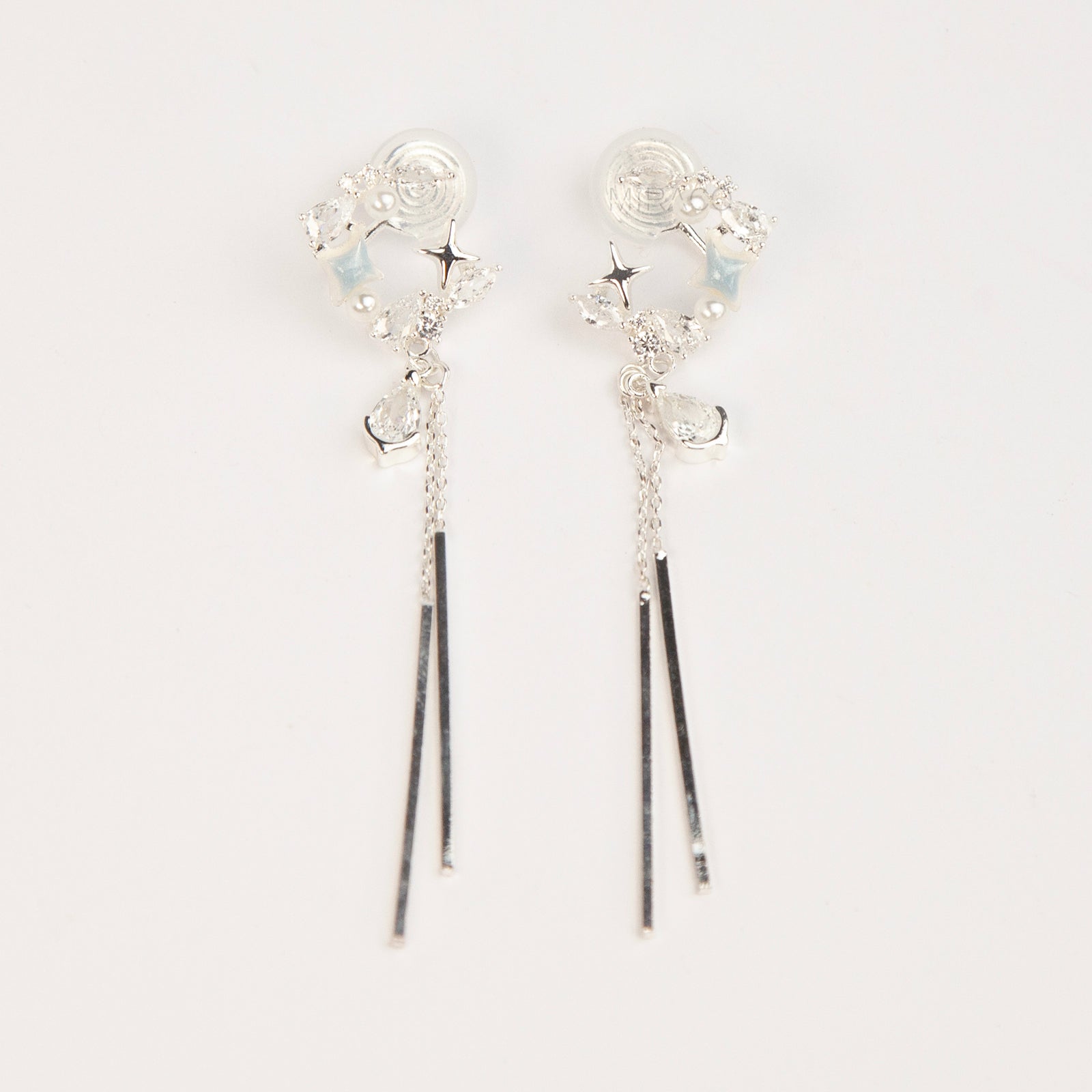 A pair of elegant silver celestial dangle clip-on earrings, featuring a crescent moon and star design accented with marquise crystals and tiny pearls, cascading into a teardrop gemstone and sleek, vertical metallic bar tassels on comfortable coil backs.