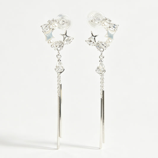A pair of elegant silver celestial dangle clip-on earrings, featuring a crescent moon and star design accented with marquise crystals and tiny pearls, cascading into a teardrop gemstone and sleek, vertical metallic bar tassels on comfortable coil backs.