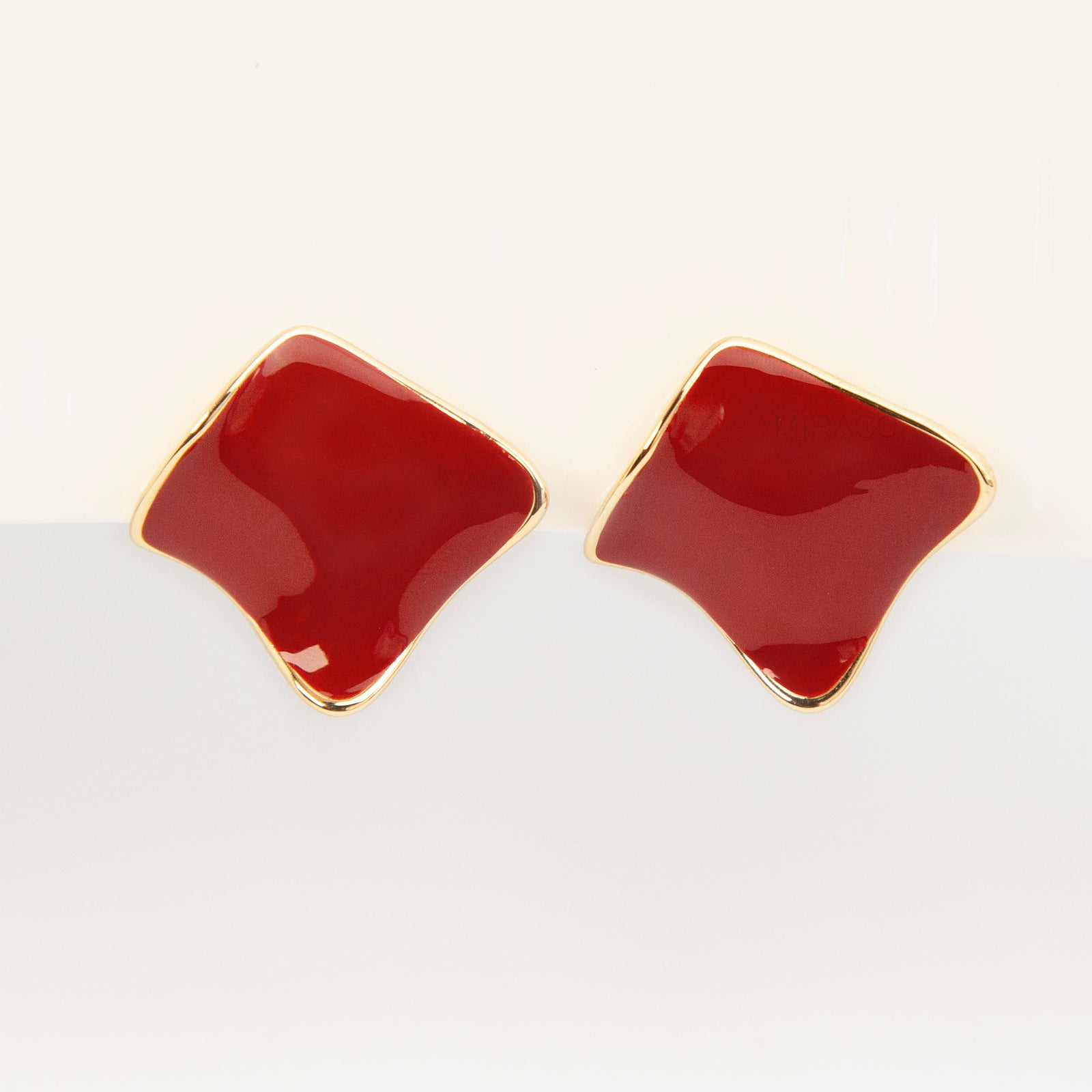 A pair of vintage-style gold clip-on earrings, showcasing a distinct wavy rhombus shape filled with smooth, glossy red enamel, fitted with comfortable, invisible-style adjustable coil backs for unpierced ears.