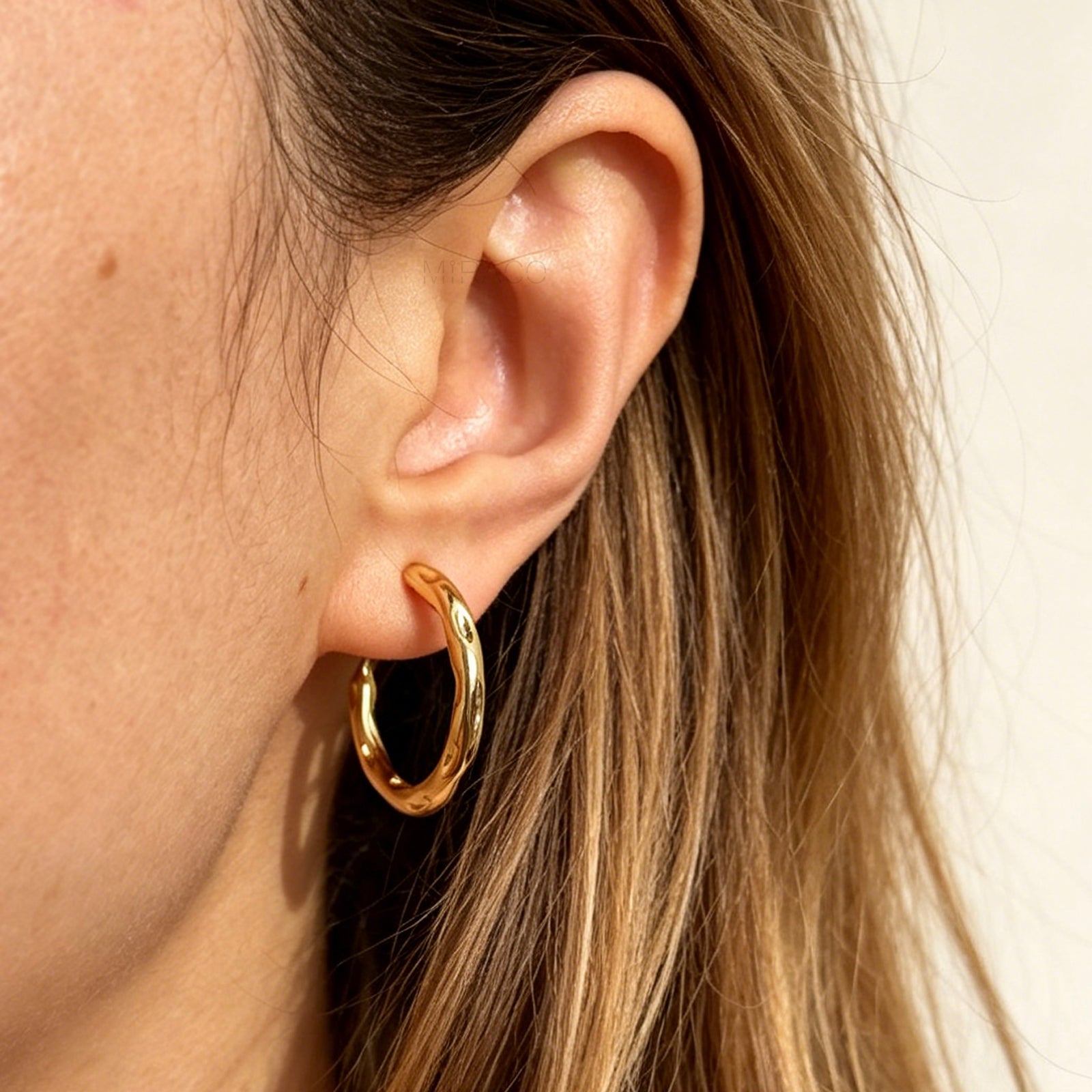A pair of gold textured, organic wavy hoop clip-on earrings, showcasing a hammered finish and comfortable coil backs with clear silicone cushions.