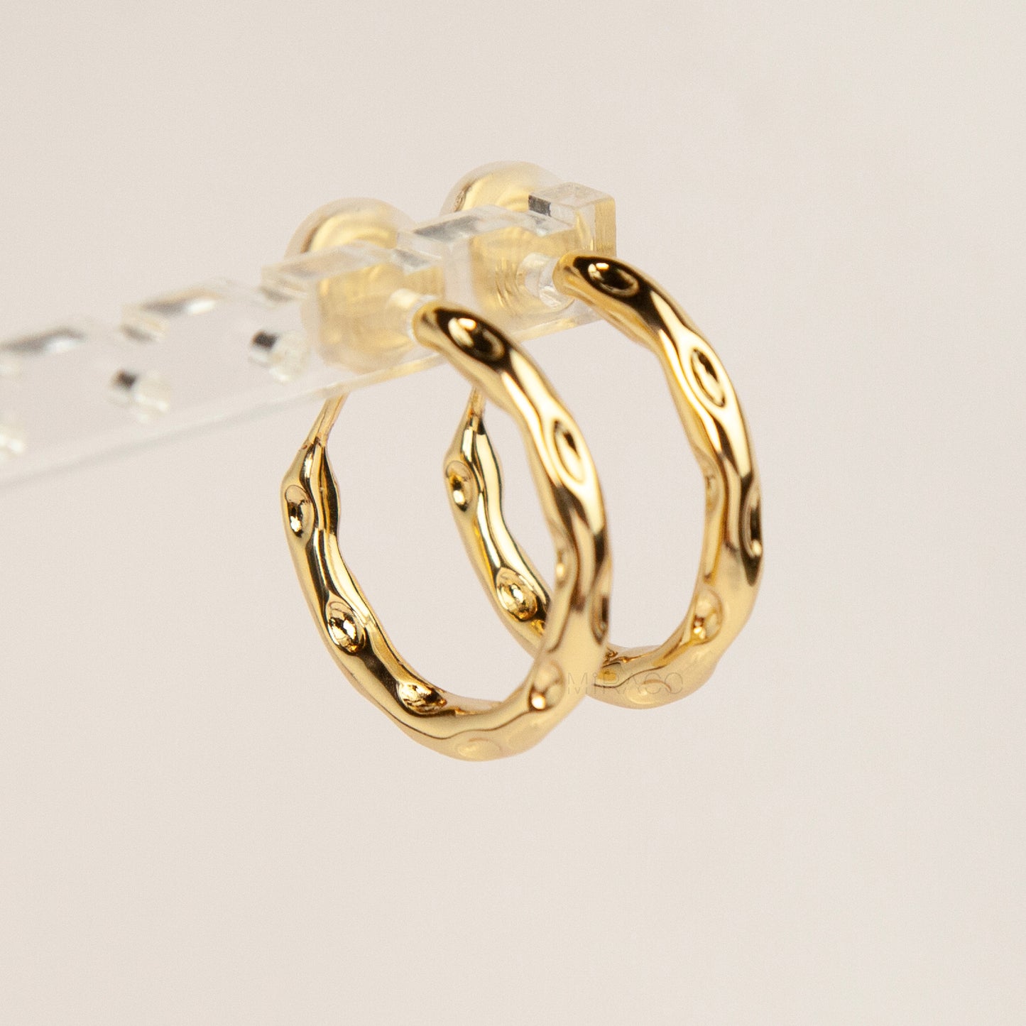 A pair of gold textured, organic wavy hoop clip-on earrings, showcasing a hammered finish and comfortable coil backs with clear silicone cushions.