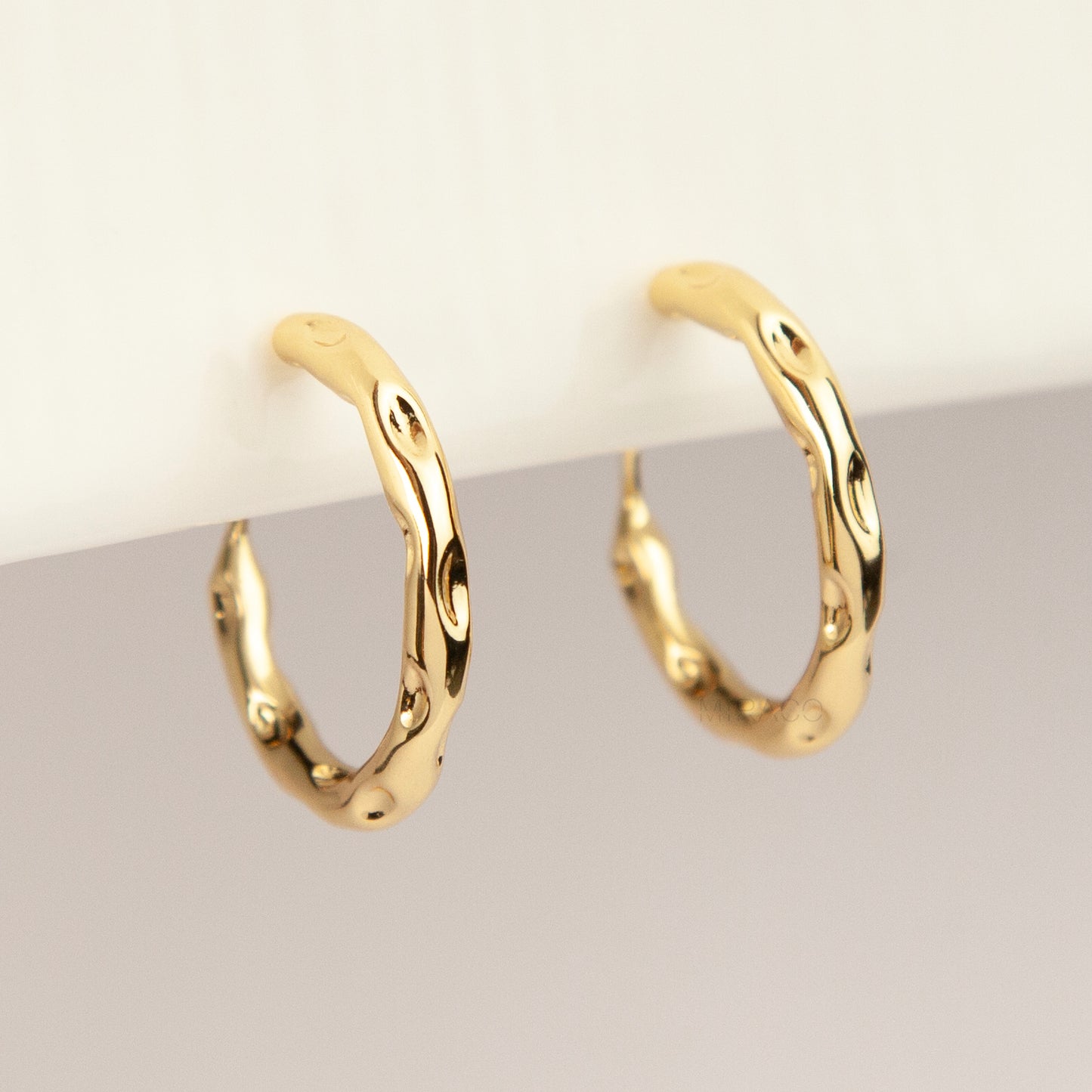 A pair of gold textured, organic wavy hoop clip-on earrings, showcasing a hammered finish and comfortable coil backs with clear silicone cushions.