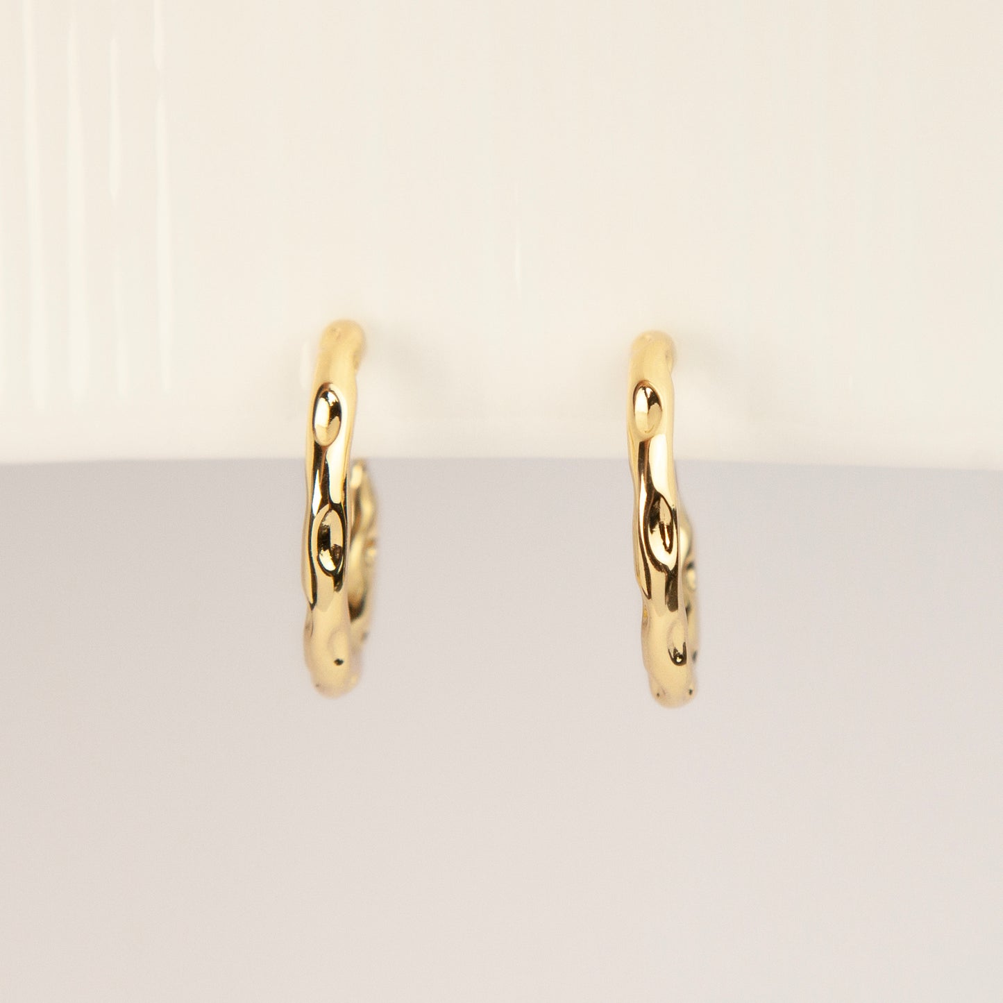 A pair of gold textured, organic wavy hoop clip-on earrings, showcasing a hammered finish and comfortable coil backs with clear silicone cushions.