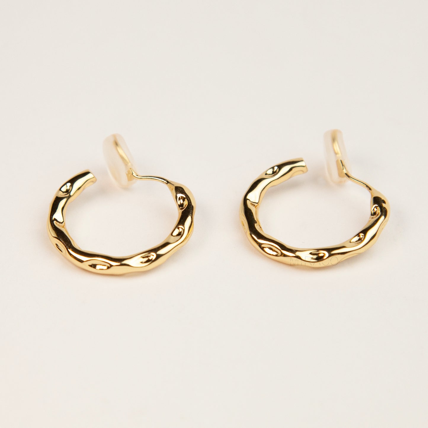 A pair of gold textured, organic wavy hoop clip-on earrings, showcasing a hammered finish and comfortable coil backs with clear silicone cushions.