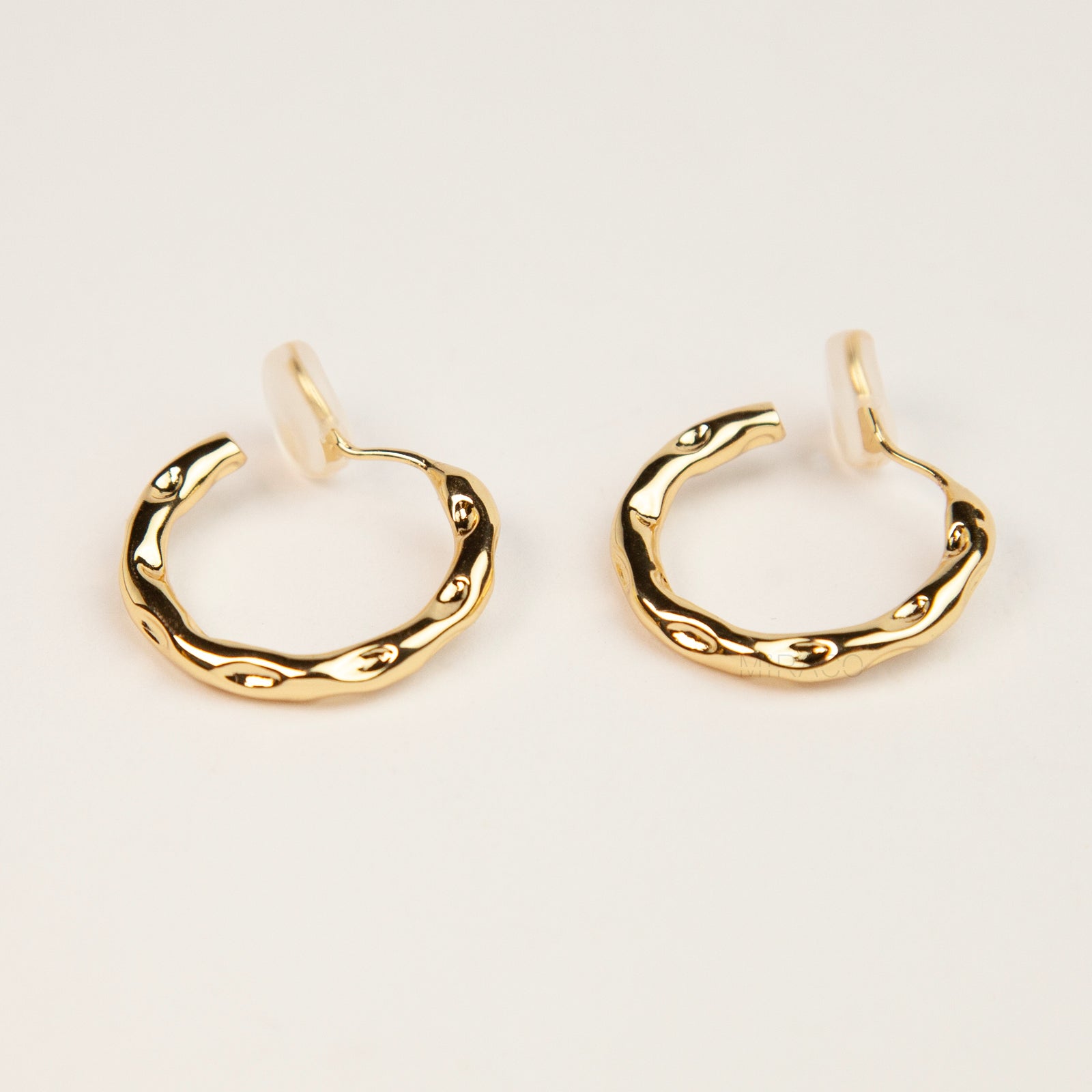 A pair of gold textured, organic wavy hoop clip-on earrings, showcasing a hammered finish and comfortable coil backs with clear silicone cushions.