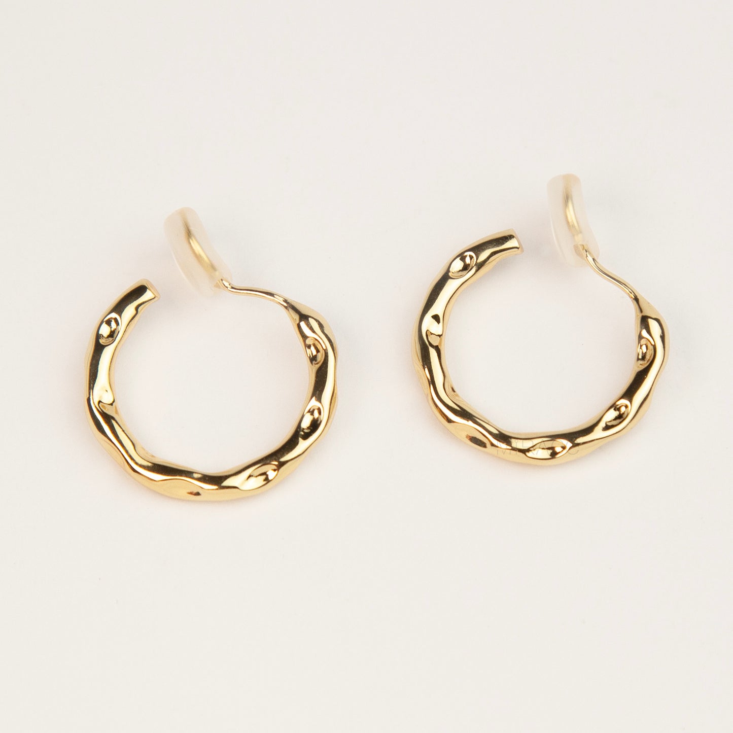 A pair of gold textured, organic wavy hoop clip-on earrings, showcasing a hammered finish and comfortable coil backs with clear silicone cushions.