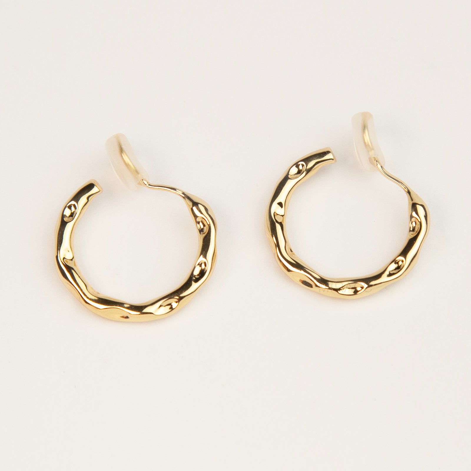 A pair of gold textured, organic wavy hoop clip-on earrings, showcasing a hammered finish and comfortable coil backs with clear silicone cushions.