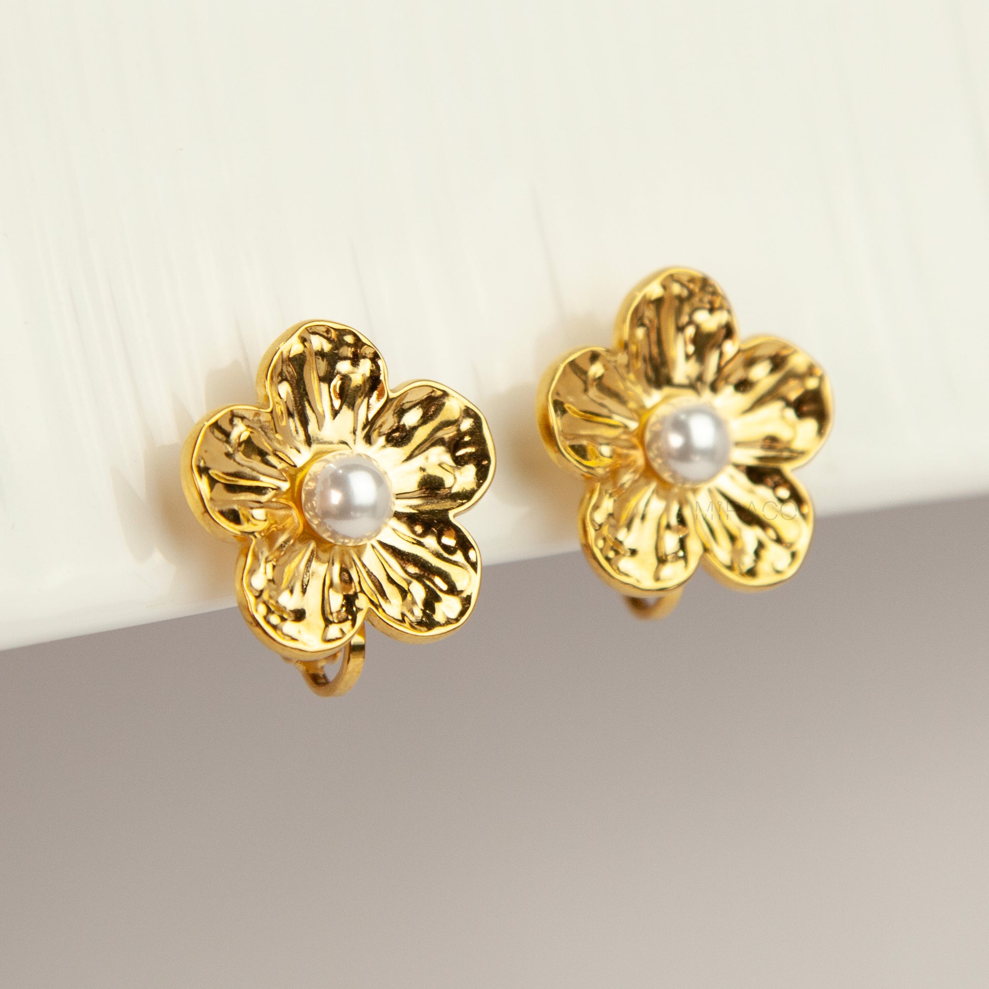 Pair of gold-toned non-pierced flower clip-on stud earrings with textured petals, white faux pearl center, and paddle back clip mechanism.