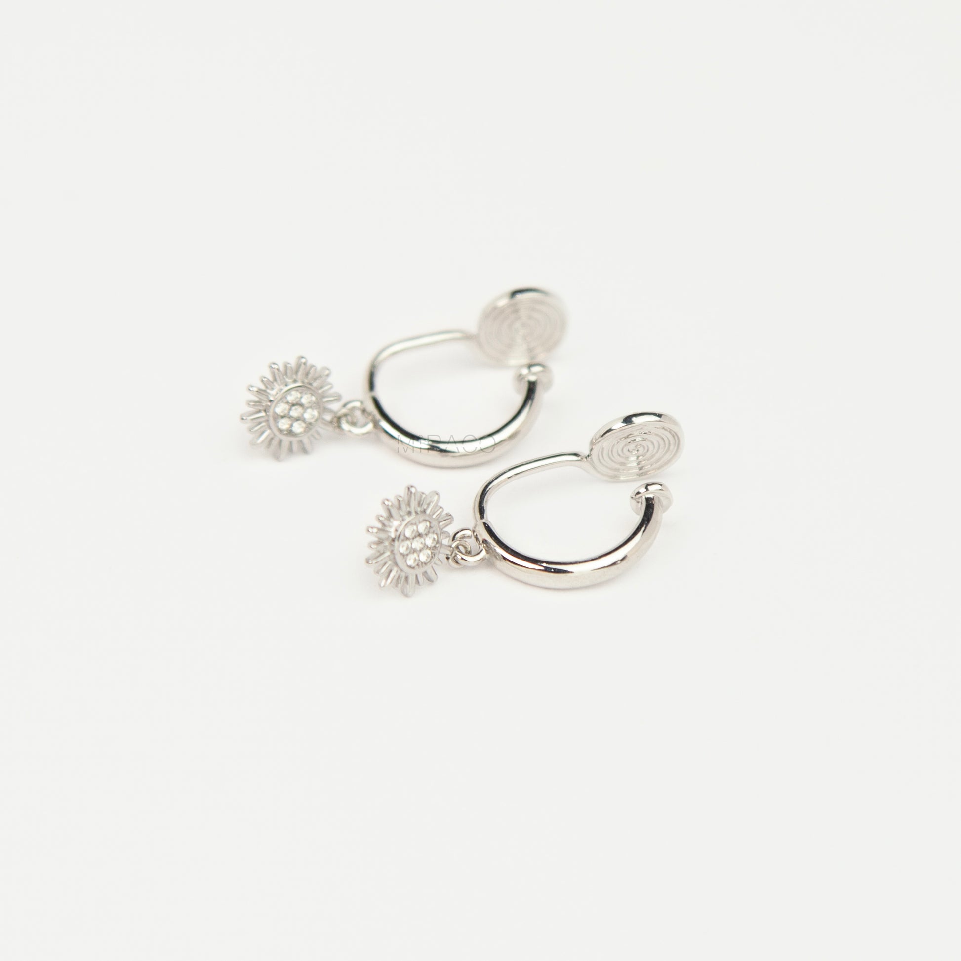 Pair of silver-toned non-pierced clip-on hoop earrings featuring dangling pave crystal sunburst charms and silicone-padded spiral coil backs.