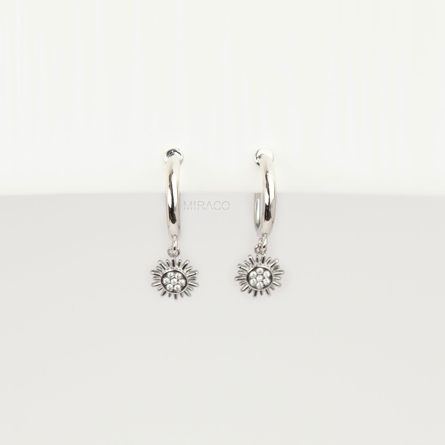 Pair of silver-toned non-pierced clip-on hoop earrings featuring dangling pave crystal sunburst charms and silicone-padded spiral coil backs.