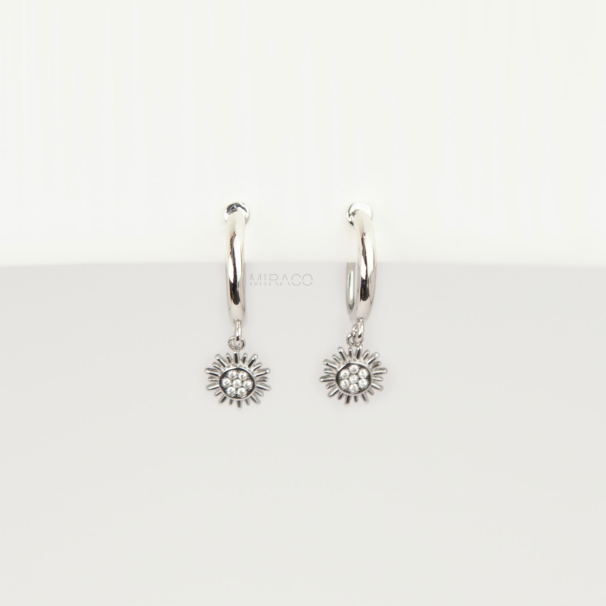 Pair of silver-toned non-pierced clip-on hoop earrings featuring dangling pave crystal sunburst charms and silicone-padded spiral coil backs.