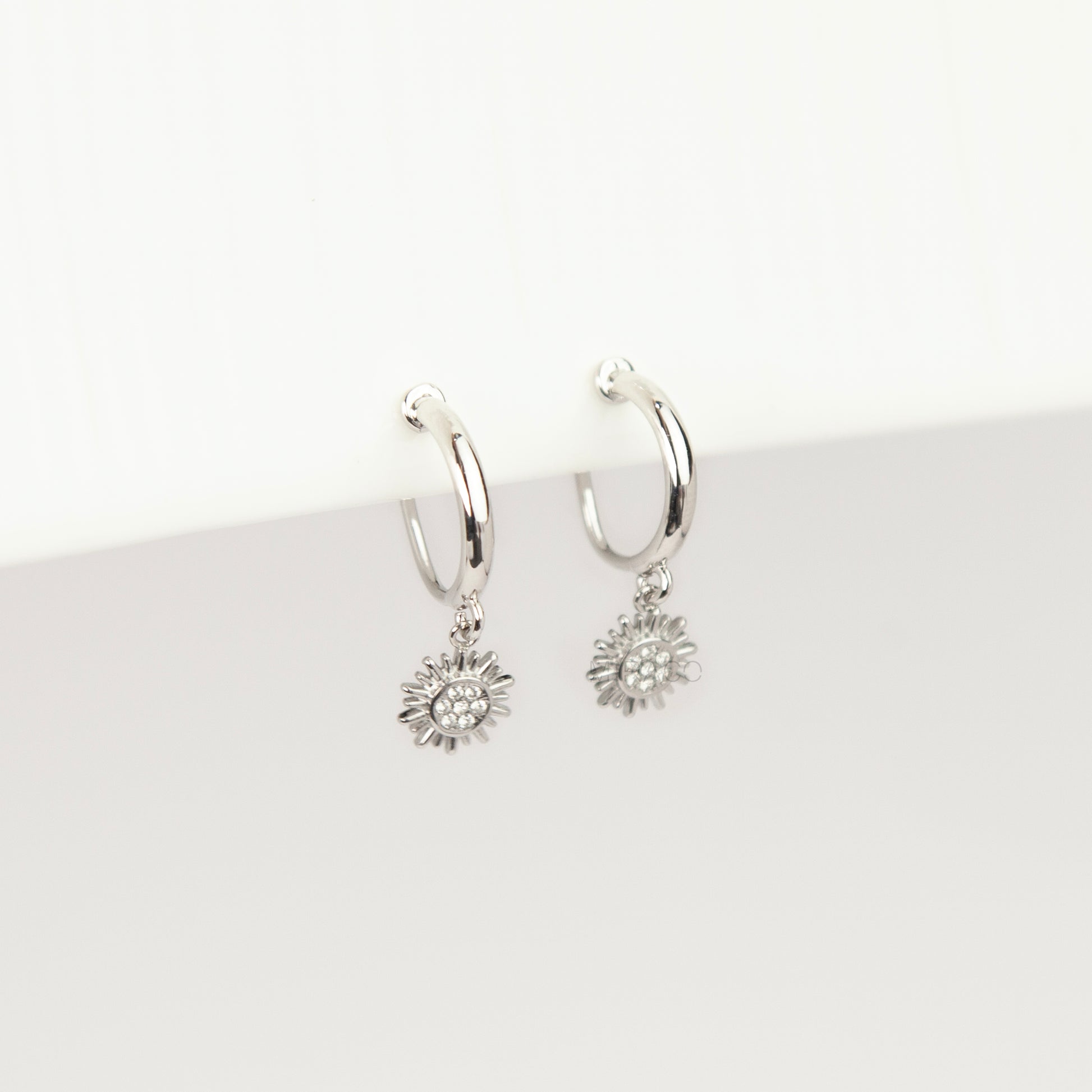 Pair of silver-toned non-pierced clip-on hoop earrings featuring dangling pave crystal sunburst charms and silicone-padded spiral coil backs.