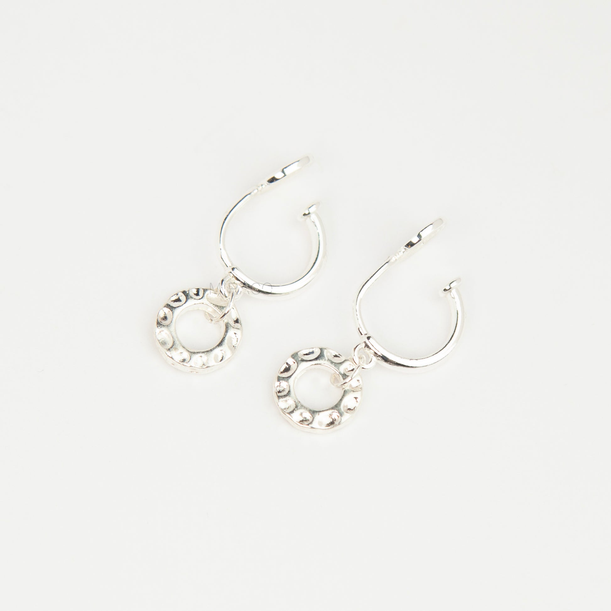 Pair of silver-toned non-pierced clip-on hoop earrings with dangling textured open circle discs and clear silicone spiral coil backs.