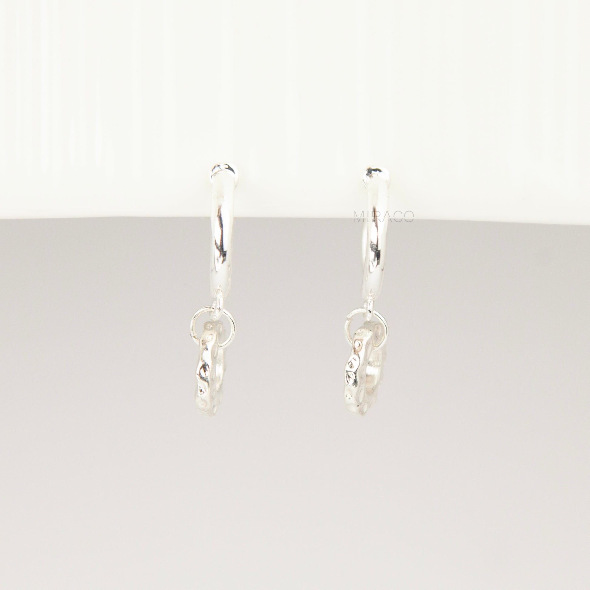 Pair of silver-toned non-pierced clip-on hoop earrings with dangling textured open circle discs and clear silicone spiral coil backs.