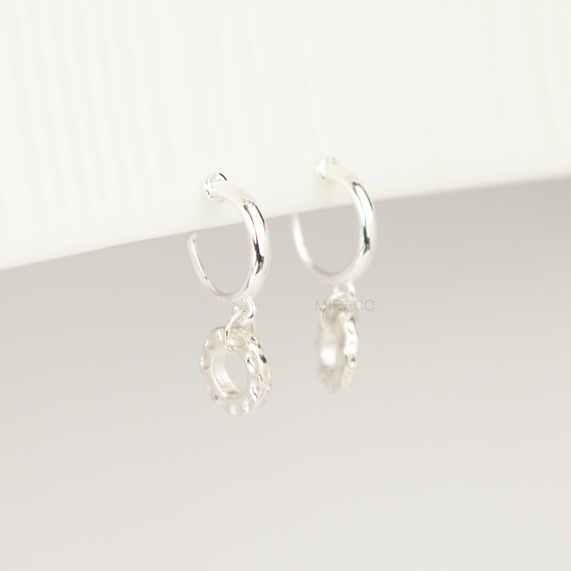 Pair of silver-toned non-pierced clip-on hoop earrings with dangling textured open circle discs and clear silicone spiral coil backs.