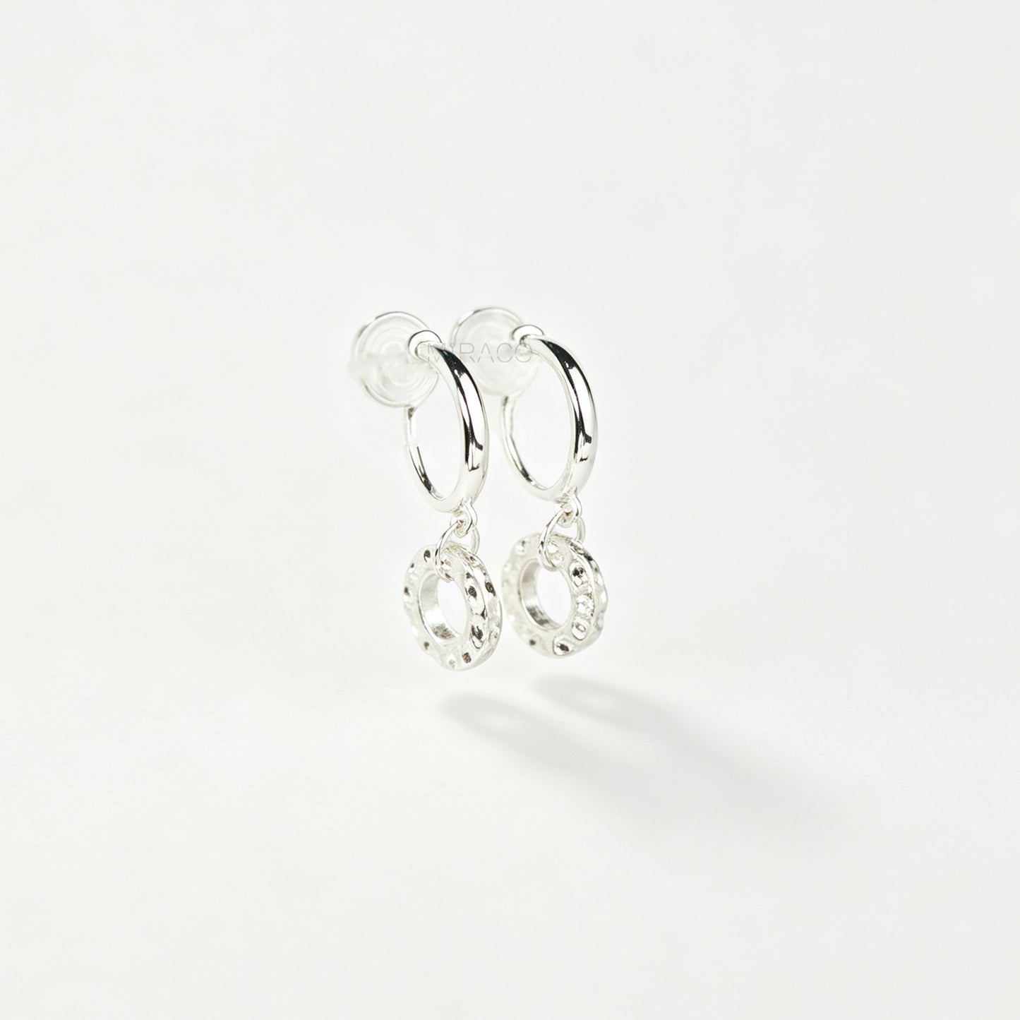 Pair of silver-toned non-pierced clip-on hoop earrings with dangling textured open circle discs and clear silicone spiral coil backs.