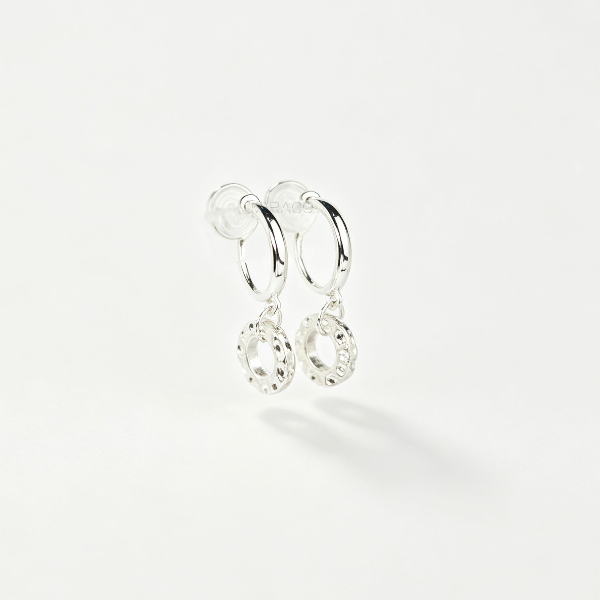 Pair of silver-toned non-pierced clip-on hoop earrings with dangling textured open circle discs and clear silicone spiral coil backs.