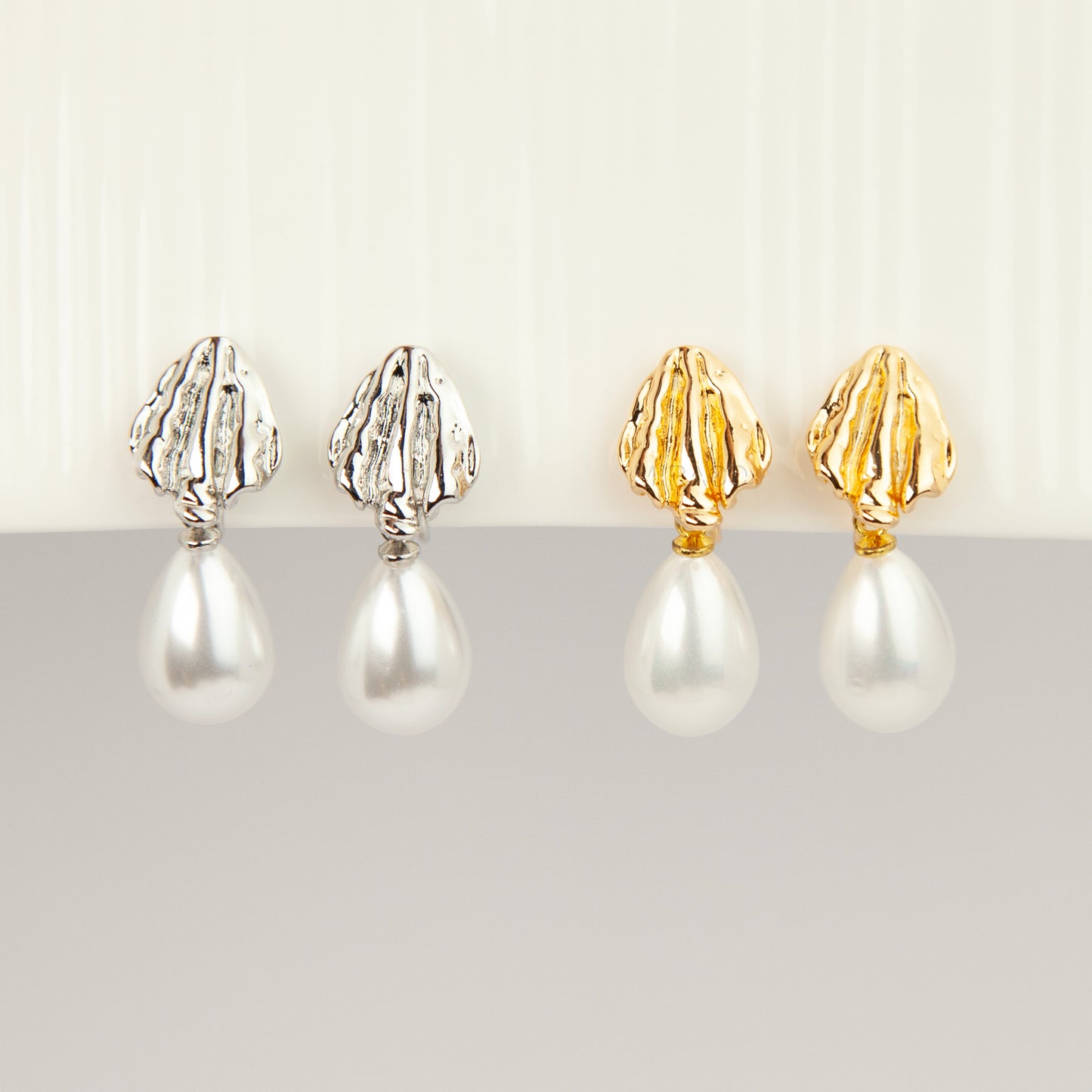 Pair of gold and silver textured shell clip-on earrings featuring white teardrop faux pearls with pain-free coil backs for non-pierced ears displayed against a clean white background.