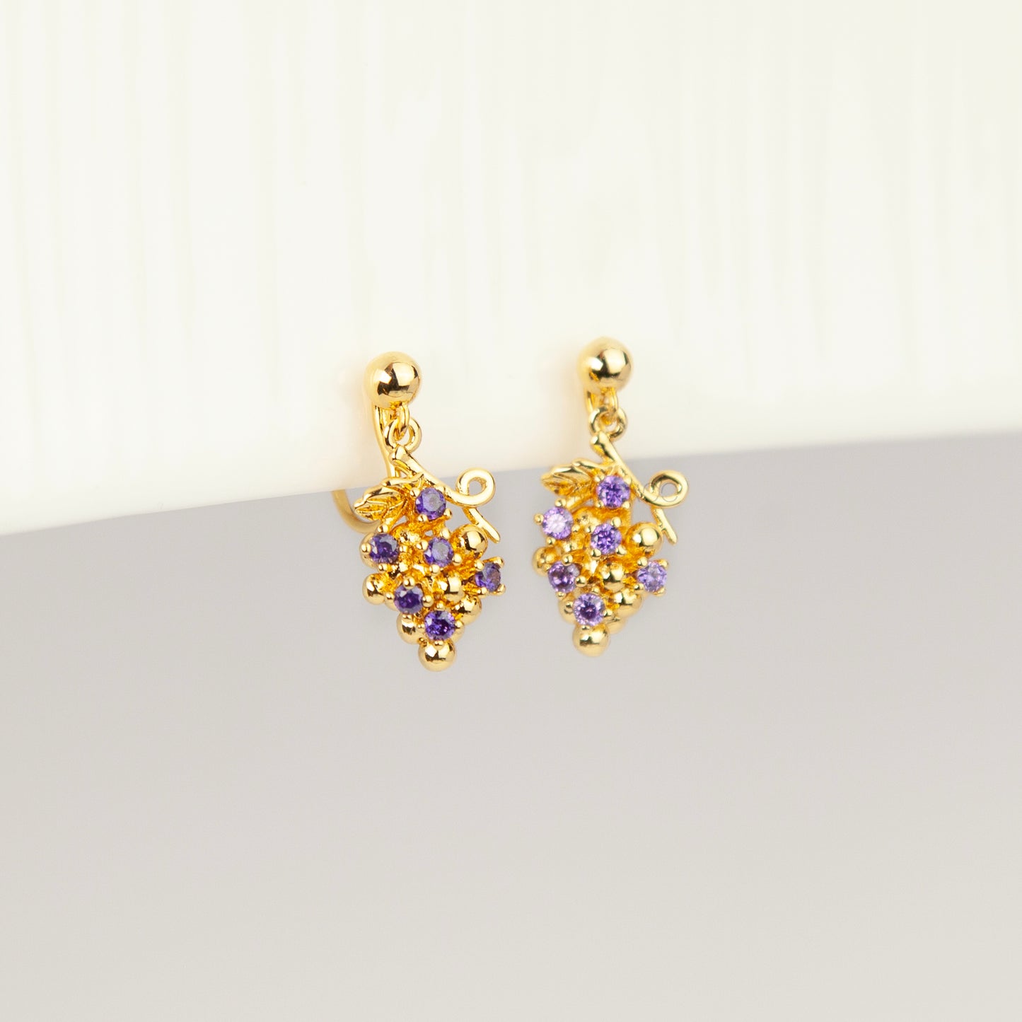 Pair of gold grape cluster clip-on drop earrings encrusted with purple crystals featuring pain-free coil backs for non-pierced ears on a white background.