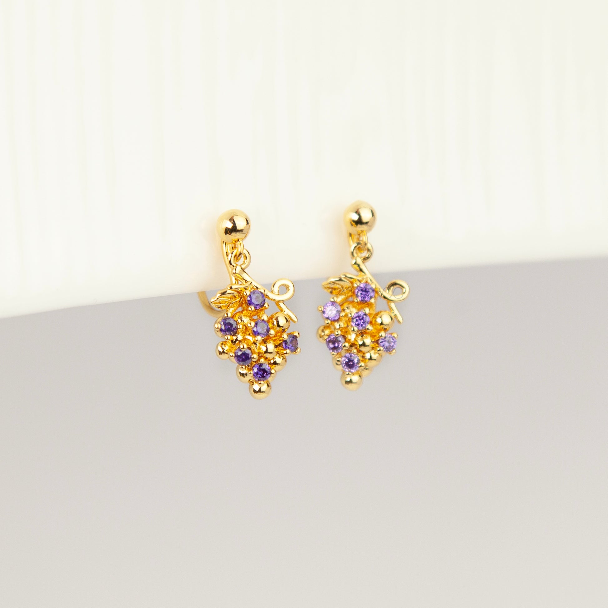 Pair of gold grape cluster clip-on drop earrings encrusted with purple crystals featuring pain-free coil backs for non-pierced ears on a white background.