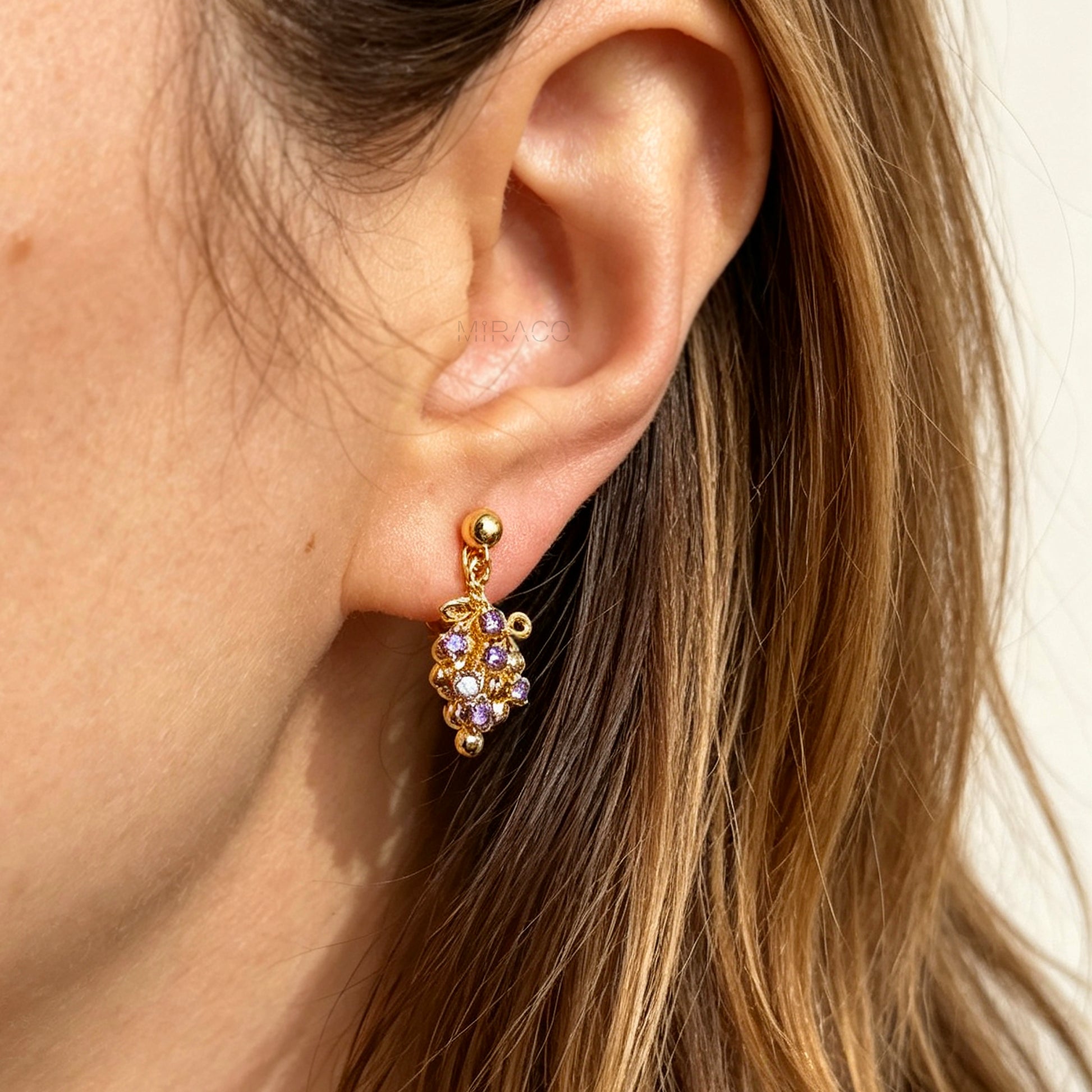 Pair of gold grape cluster clip-on drop earrings encrusted with purple crystals featuring pain-free coil backs for non-pierced ears on a white background.