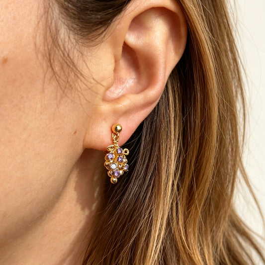 Pair of gold grape cluster clip-on drop earrings encrusted with purple crystals featuring pain-free coil backs for non-pierced ears on a white background.