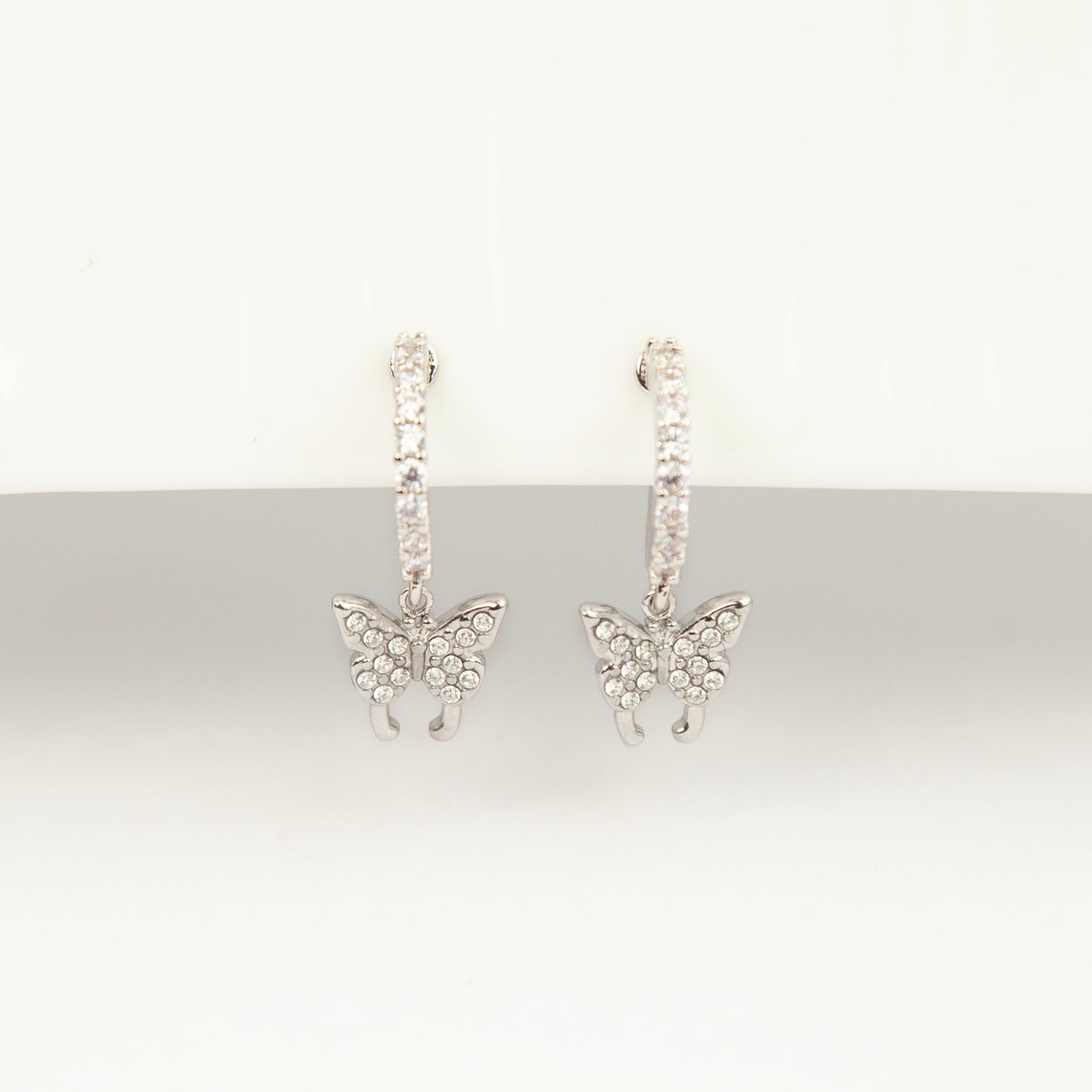 Pair of silver clip-on huggie hoop earrings featuring pavé cubic zirconia butterfly charms and pain-free coil backs for non-pierced ears against a white background.