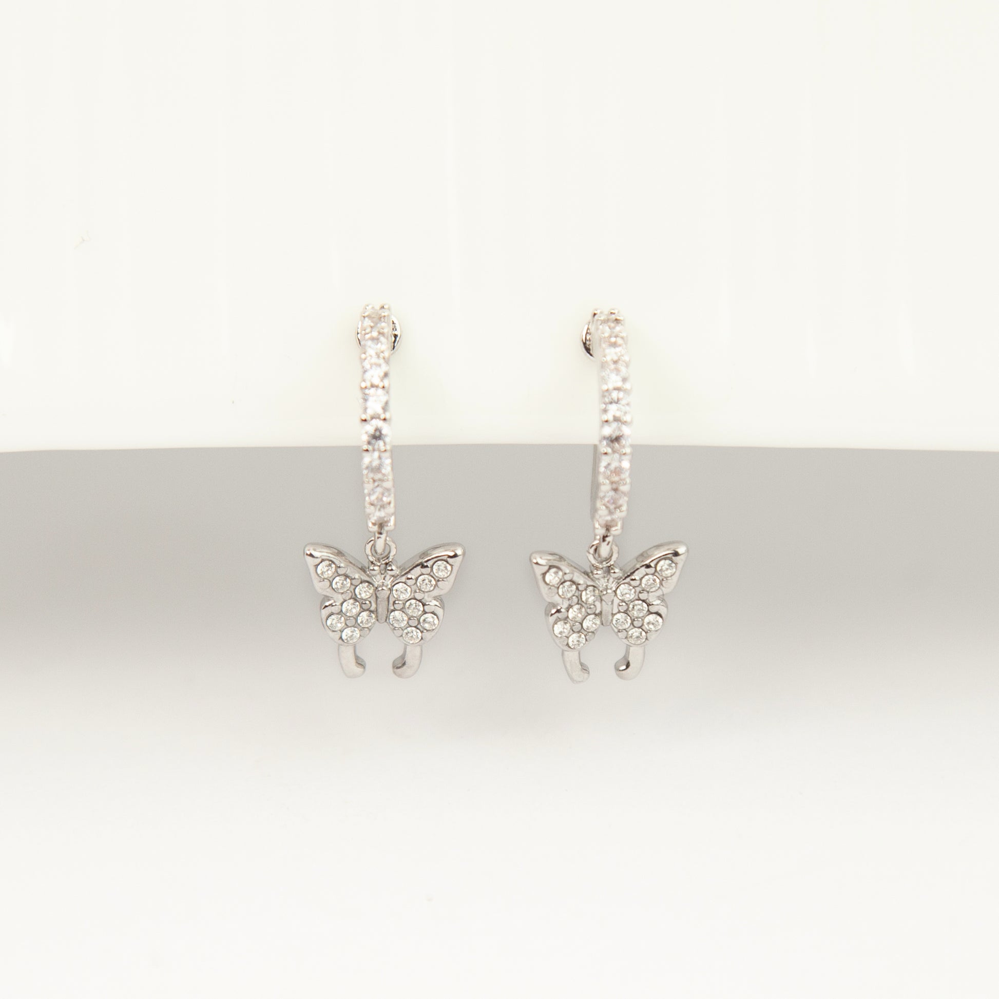 Pair of silver clip-on huggie hoop earrings featuring pavé cubic zirconia butterfly charms and pain-free coil backs for non-pierced ears against a white background.