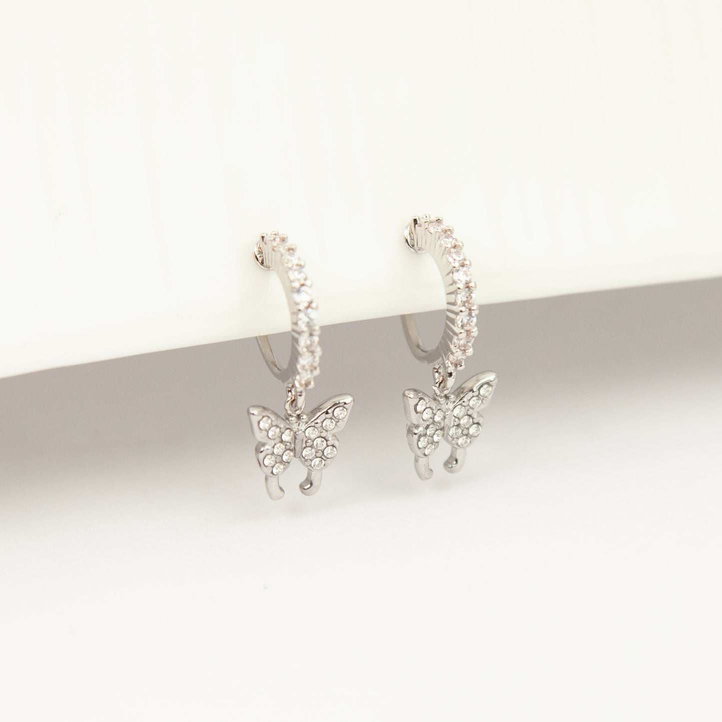 Pair of silver clip-on huggie hoop earrings featuring pavé cubic zirconia butterfly charms and pain-free coil backs for non-pierced ears against a white background.