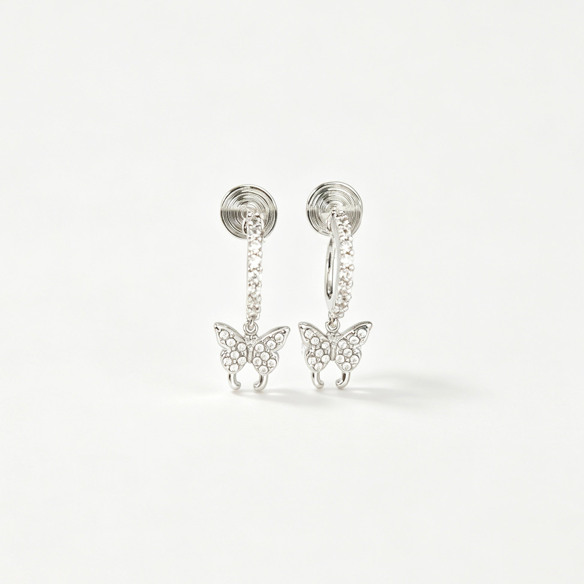 Pair of silver clip-on huggie hoop earrings featuring pavé cubic zirconia butterfly charms and pain-free coil backs for non-pierced ears against a white background.