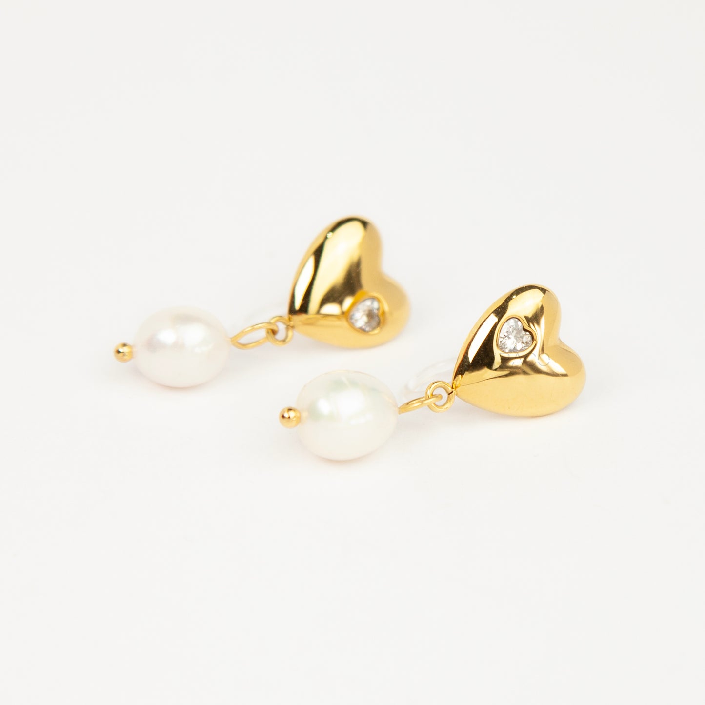 Pair of polished gold puffy heart clip-on earrings with star-set cz detail and dangling white pearls featuring clear invisible resin backs for non-pierced ears against a white backdrop.