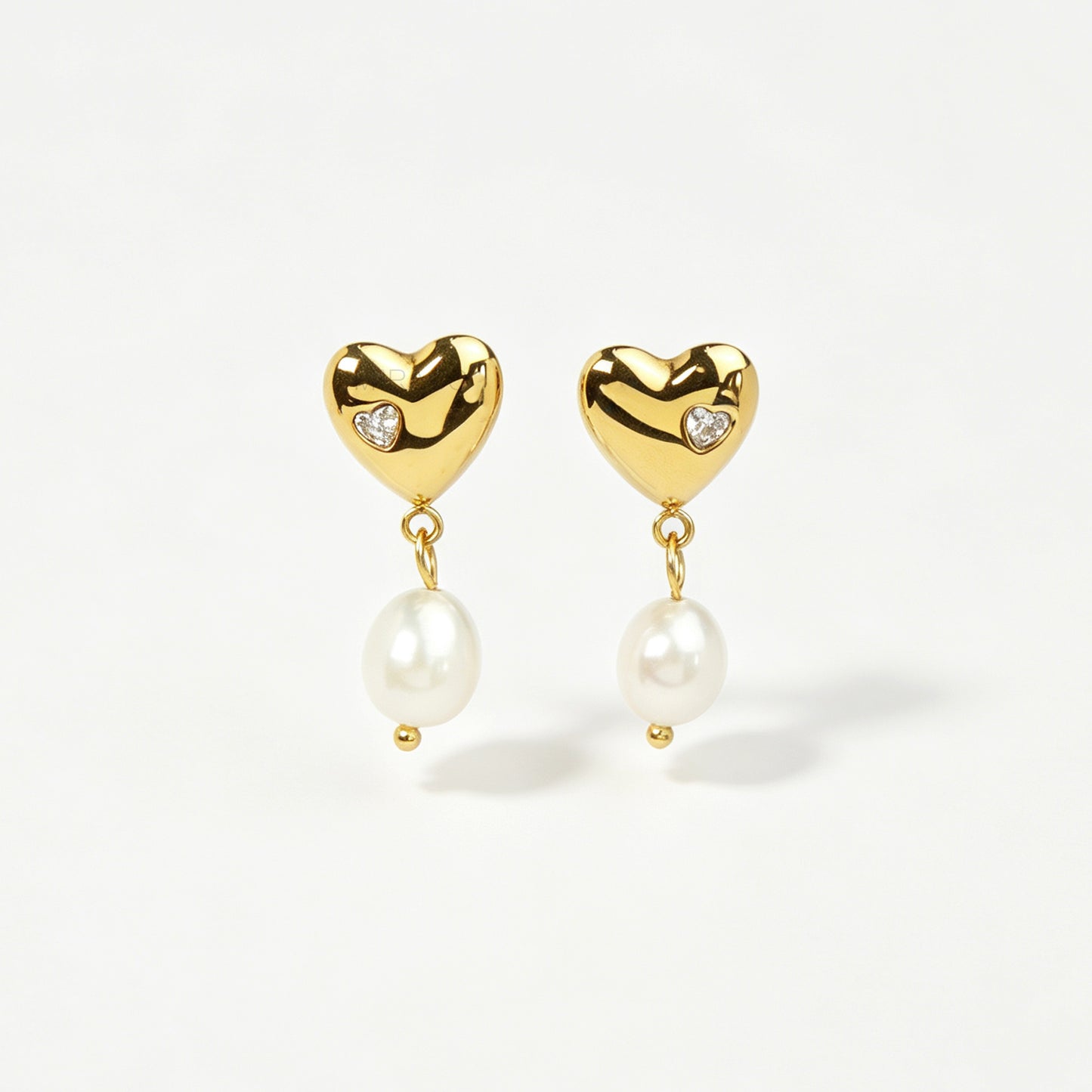 Pair of polished gold puffy heart clip-on earrings with star-set cz detail and dangling white pearls featuring clear invisible resin backs for non-pierced ears against a white backdrop.