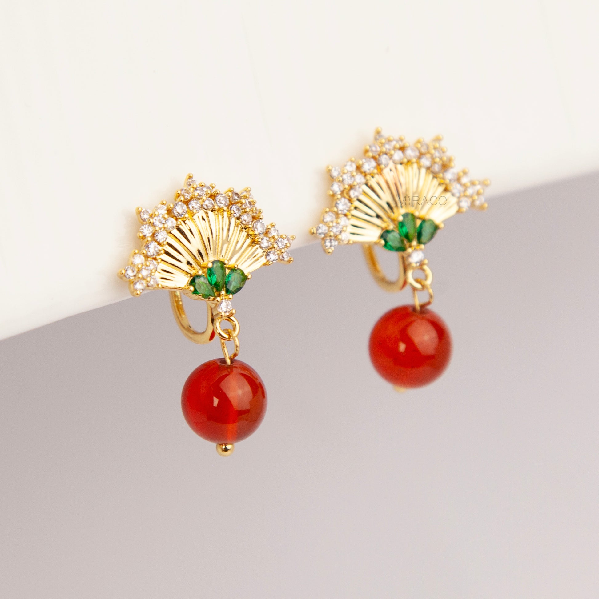 Gold Art Deco fan-shaped clip-on earrings with green crystal accents, pavé CZ trim, and hanging red agate bead drops 