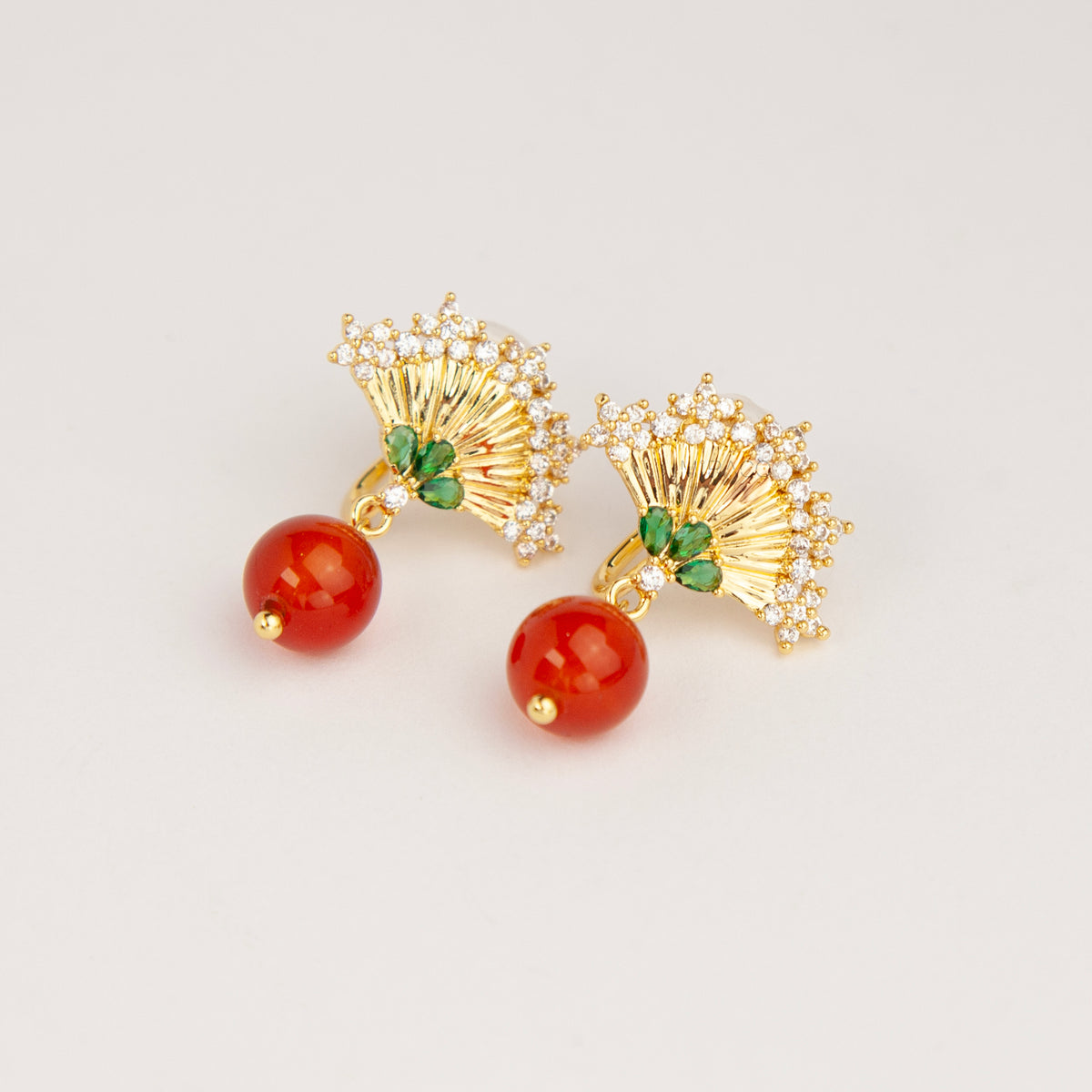 Gold Art Deco fan-shaped clip-on earrings with green crystal accents, pavé CZ trim, and hanging red agate bead drops 