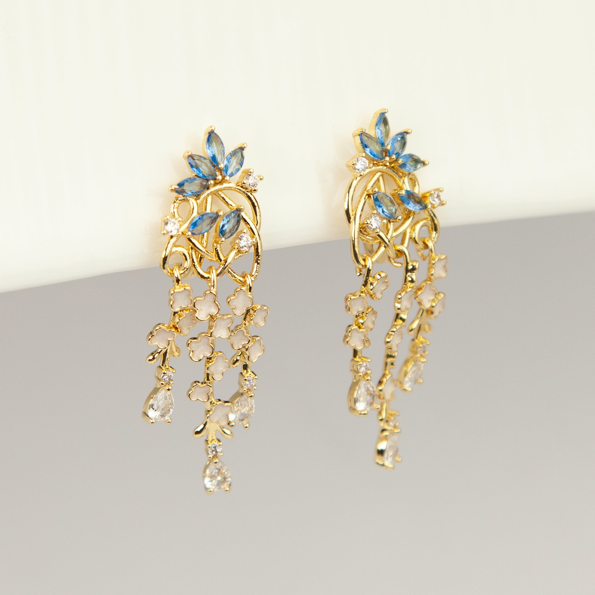 Gold coil-back clip-on chandelier earrings featuring blue marquise petals and cascading white flower dangles with teardrop crystals