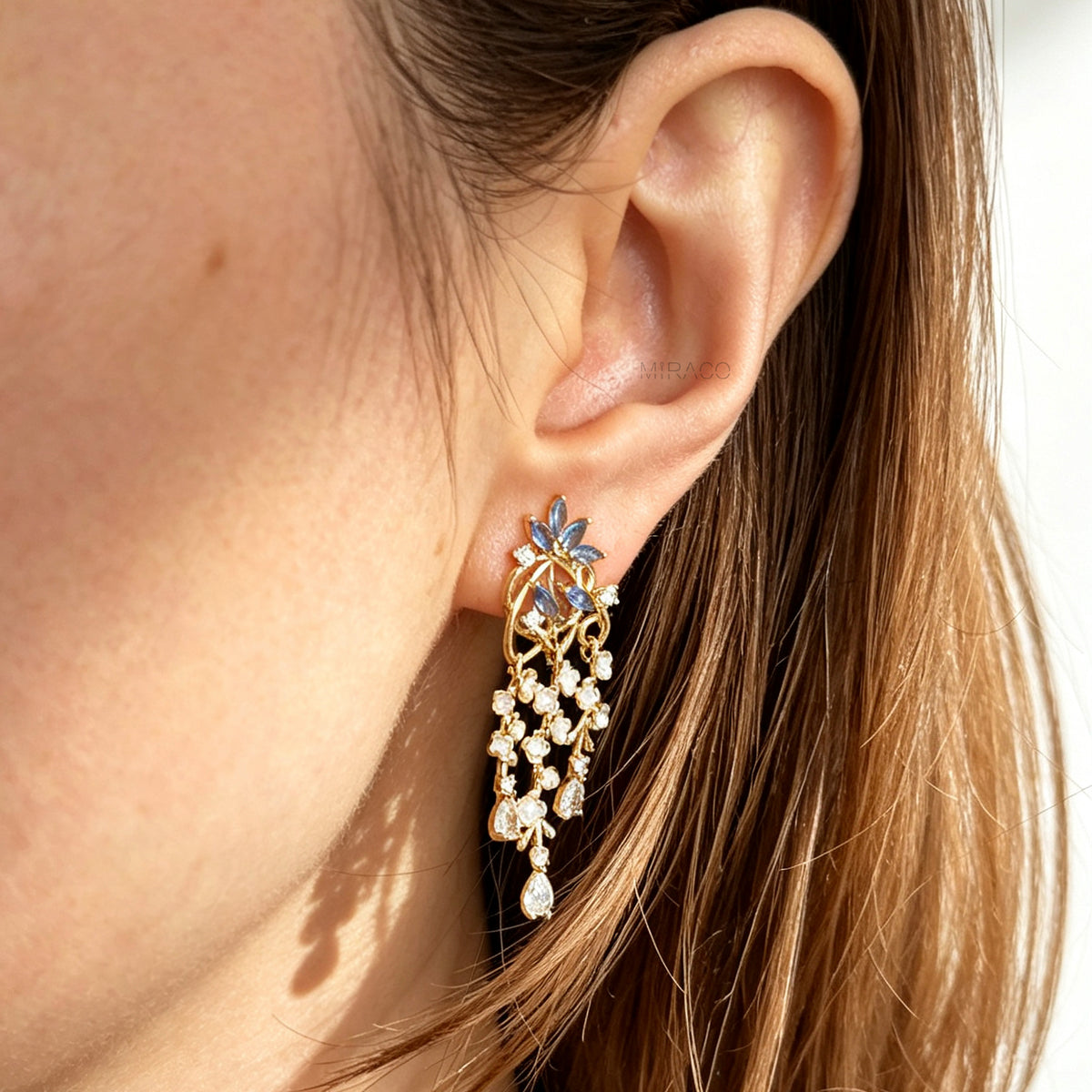 Gold coil-back clip-on chandelier earrings featuring blue marquise petals and cascading white flower dangles with teardrop crystals worn on a ear.