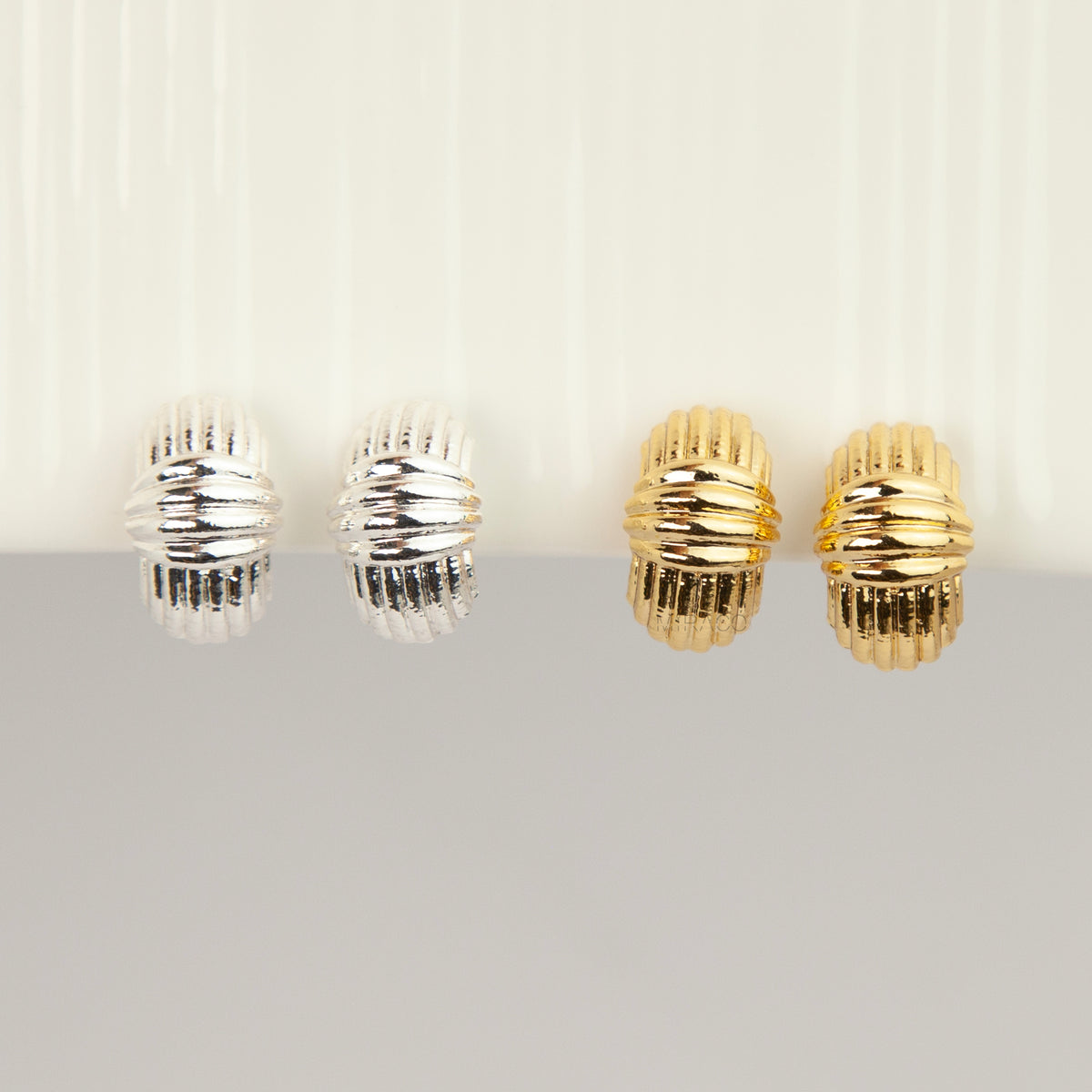 Four ribbed hoop earrings in silver and gold finishes, arranged in pairs on a light gray and white background.