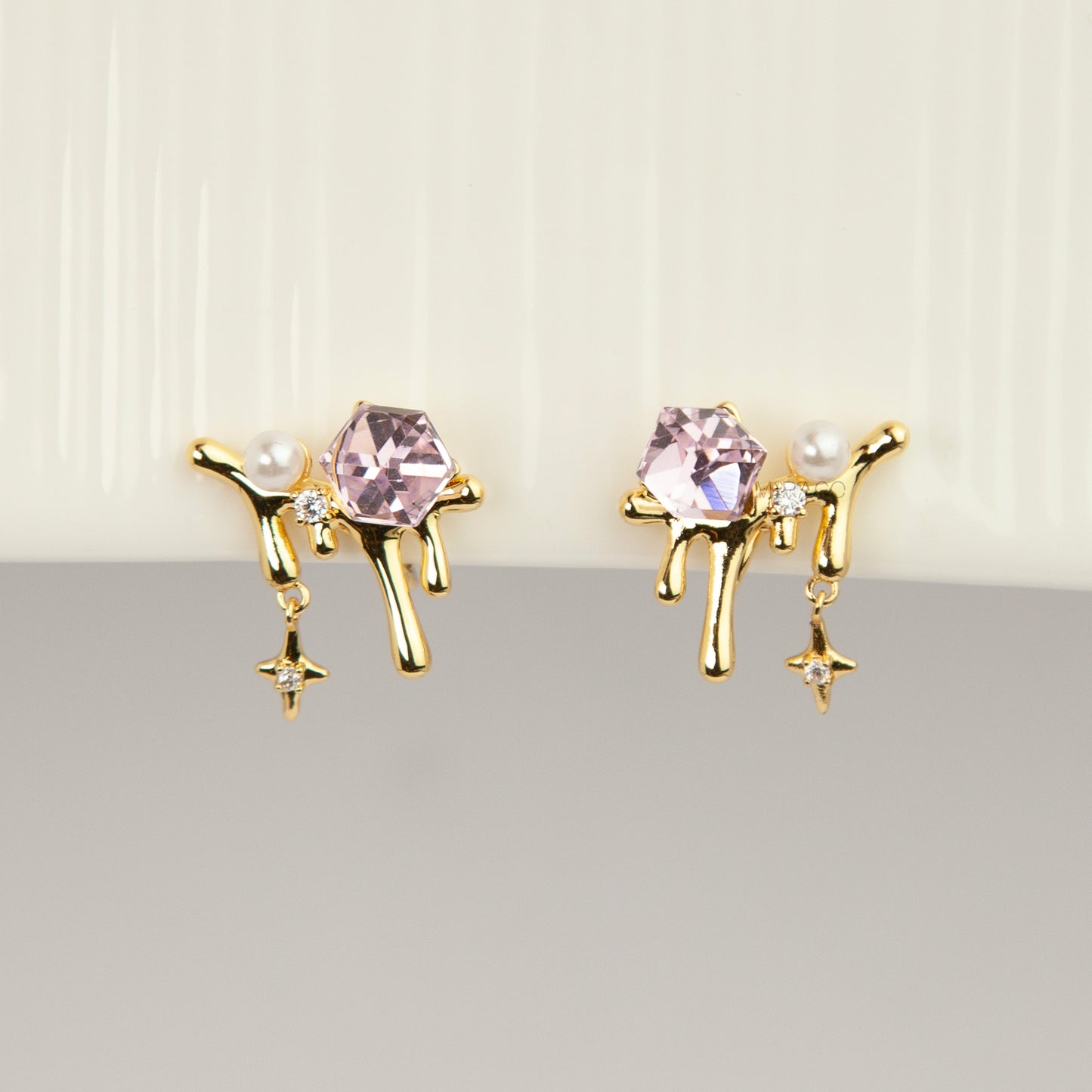 Gold coil-back clip-on earrings featuring a molten liquid drip design with a geometric pink crystal, white pearl, and dangling starburst charm.