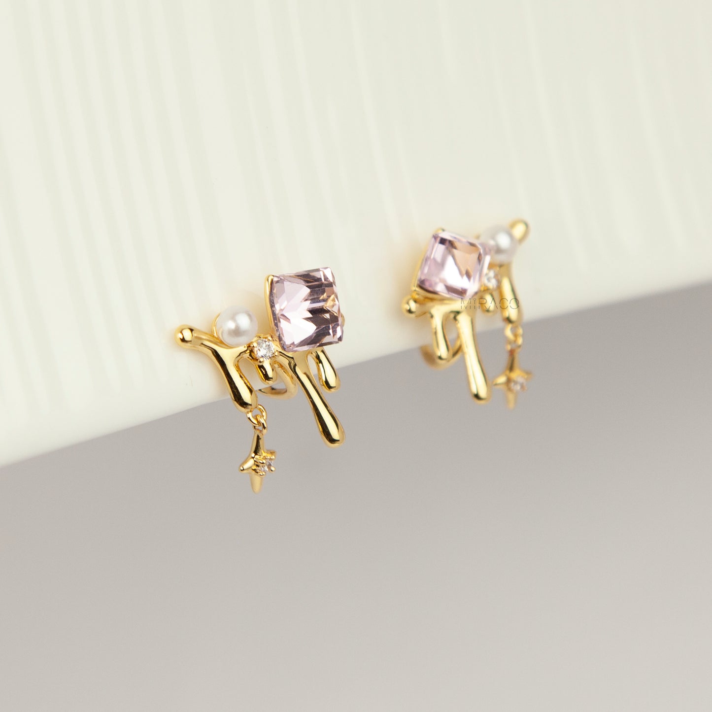 Gold coil-back clip-on earrings featuring a molten liquid drip design with a geometric pink crystal, white pearl, and dangling starburst charm.