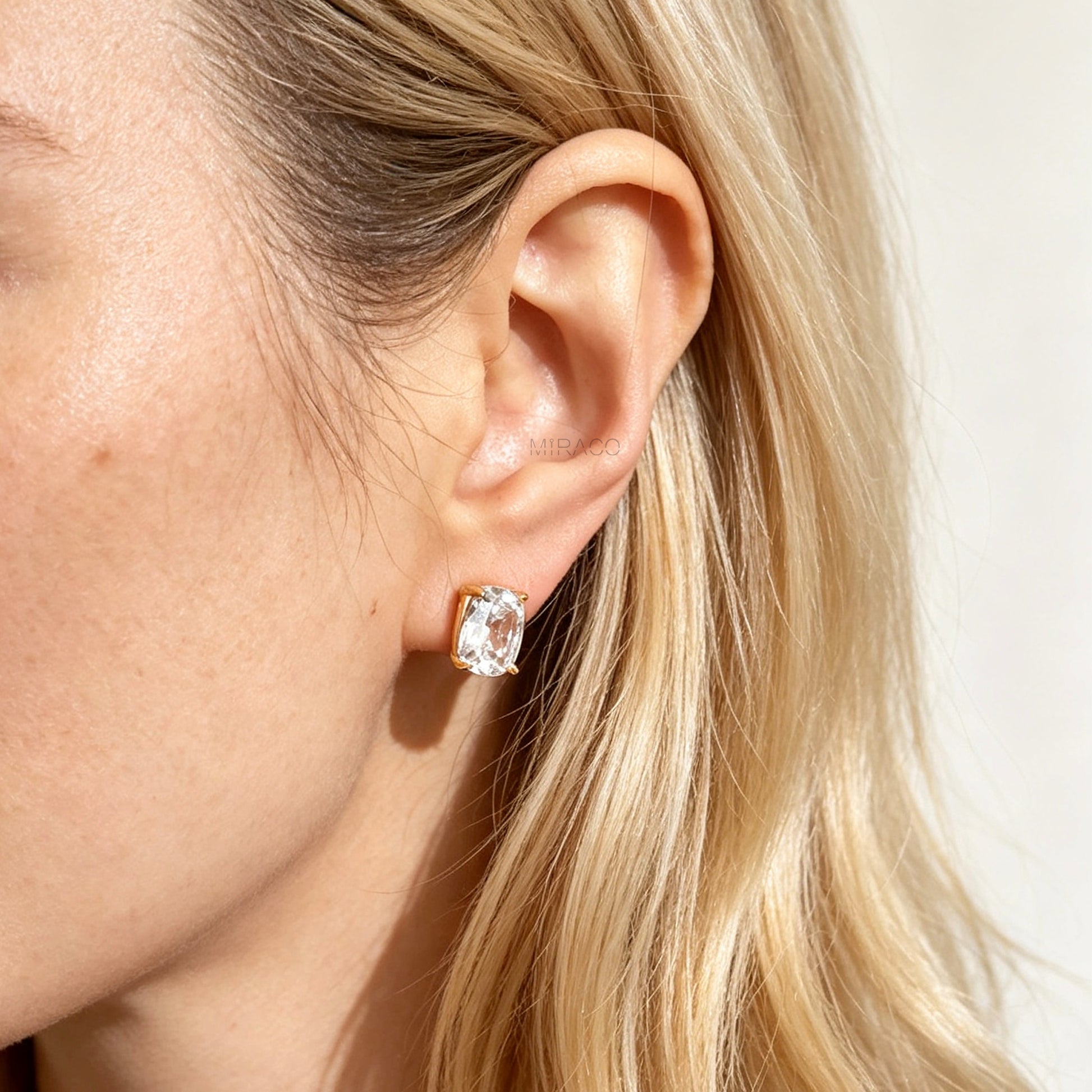 Gold-tone clip-on stud earrings set with large cushion-cut clear crystals and round silicone backs worn on a model's ear.