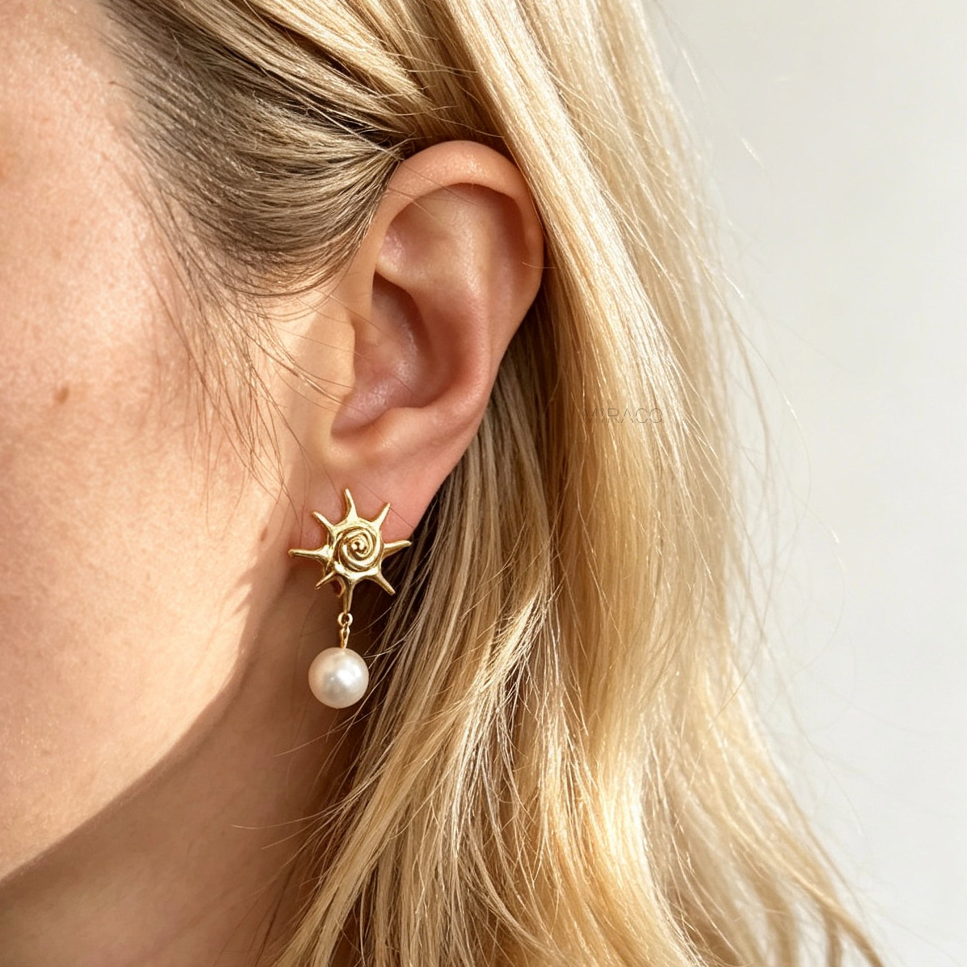 Gold coil-back clip-on earrings with a swirled sunburst star motif and a dangling white pearl worn on a model's ear.
