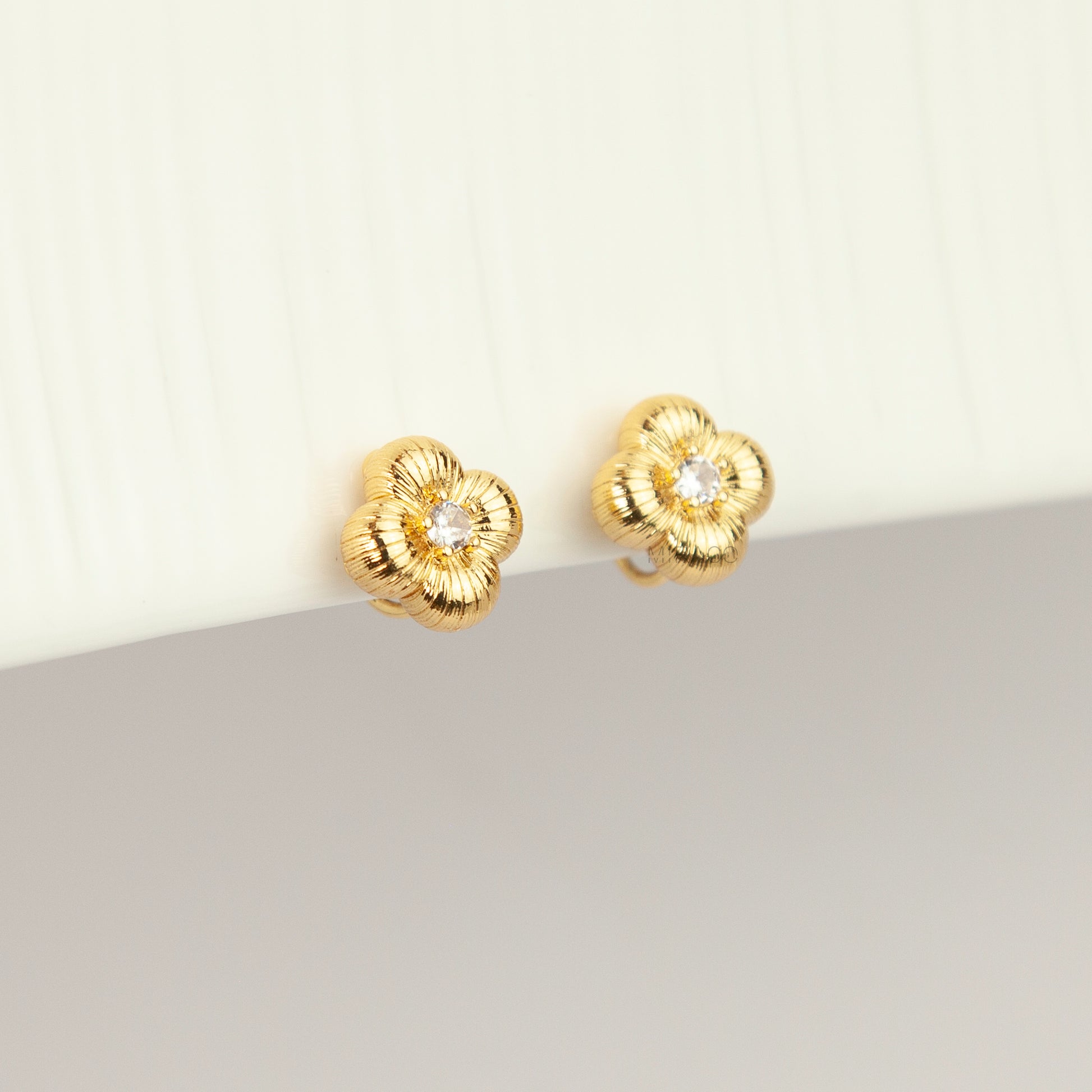 Textured gold clover-shaped floral clip-on stud earrings set with a round central crystal and a coil-back closure.