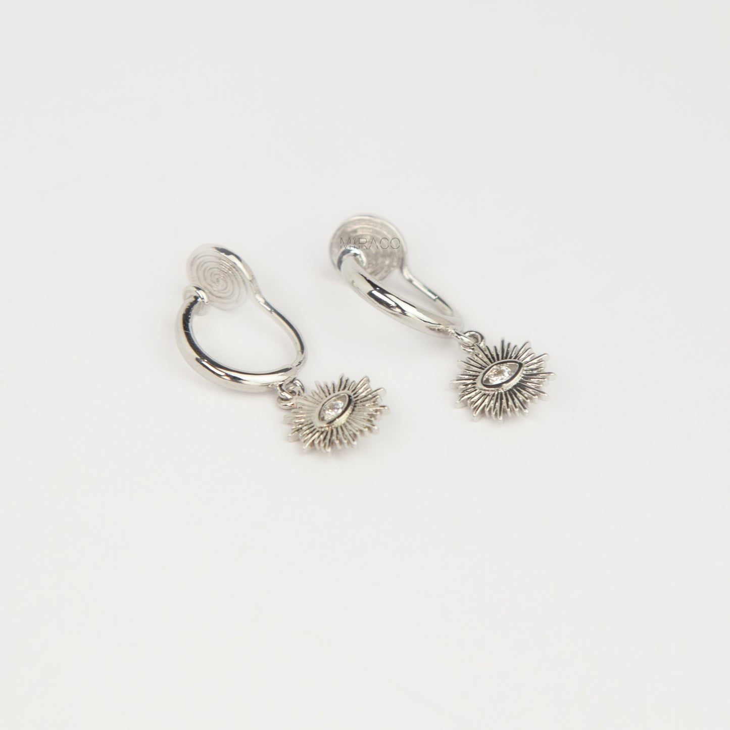 Silver coil-back clip-on hoop earrings featuring a dangling sunburst charm with a central oval crystal evil eye motif 