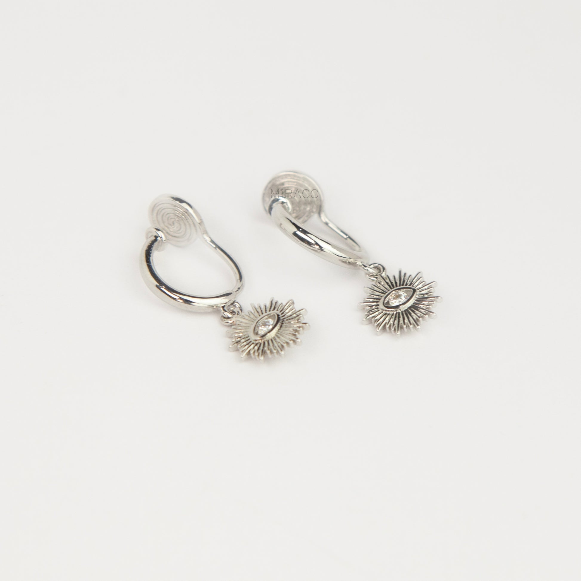 Silver coil-back clip-on hoop earrings featuring a dangling sunburst charm with a central oval crystal evil eye motif 