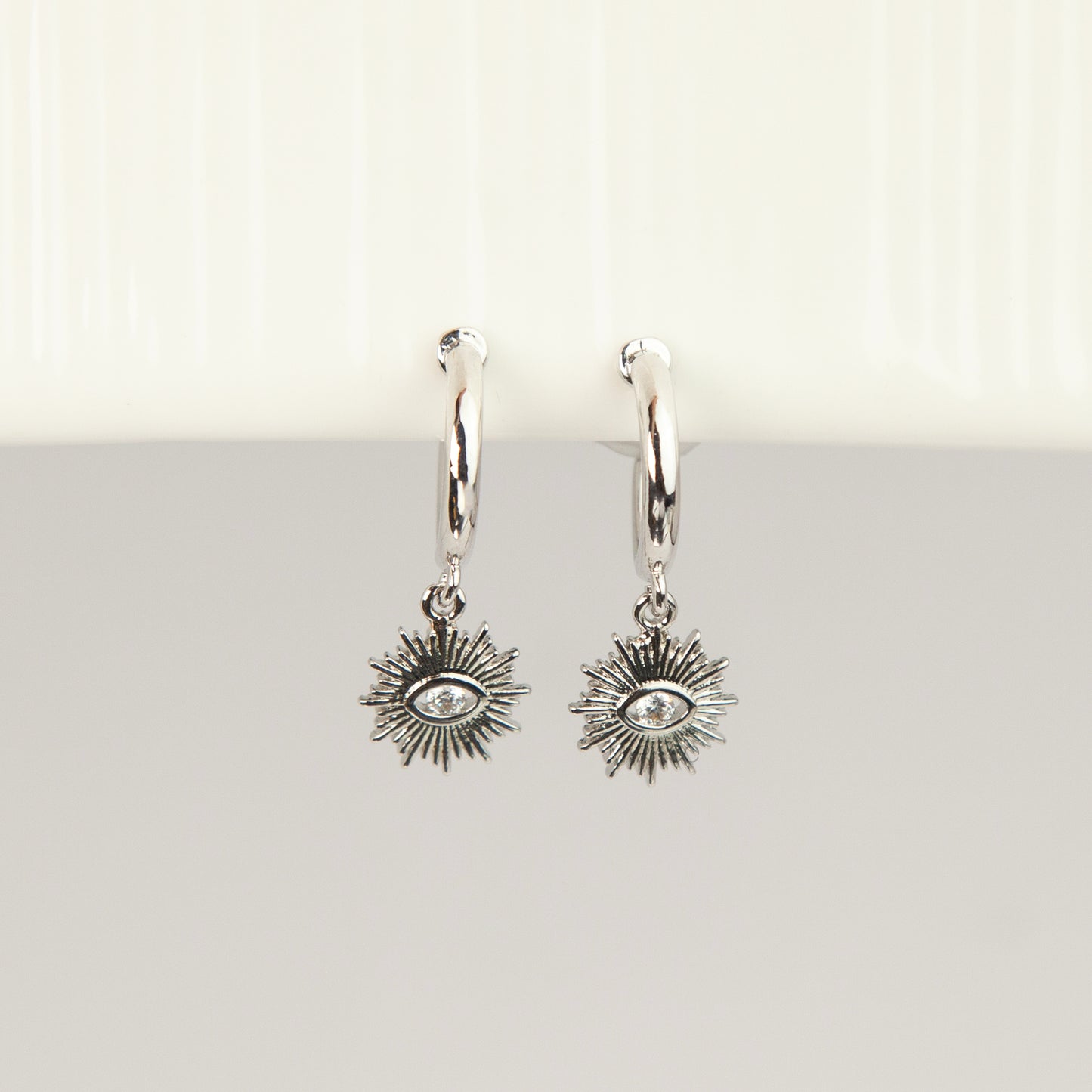 Silver coil-back clip-on hoop earrings featuring a dangling sunburst charm with a central oval crystal evil eye motif 