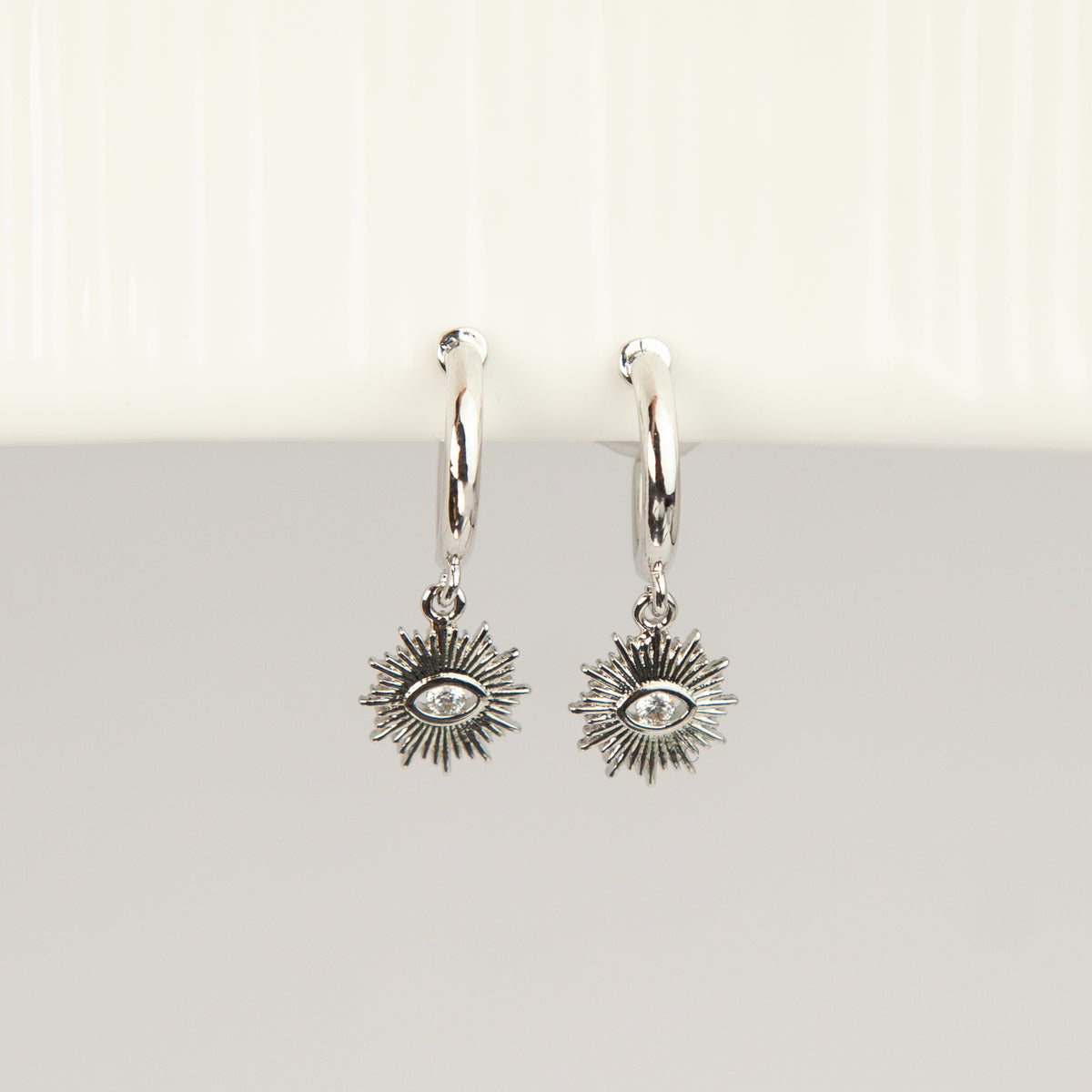 Silver coil-back clip-on hoop earrings featuring a dangling sunburst charm with a central oval crystal evil eye motif 