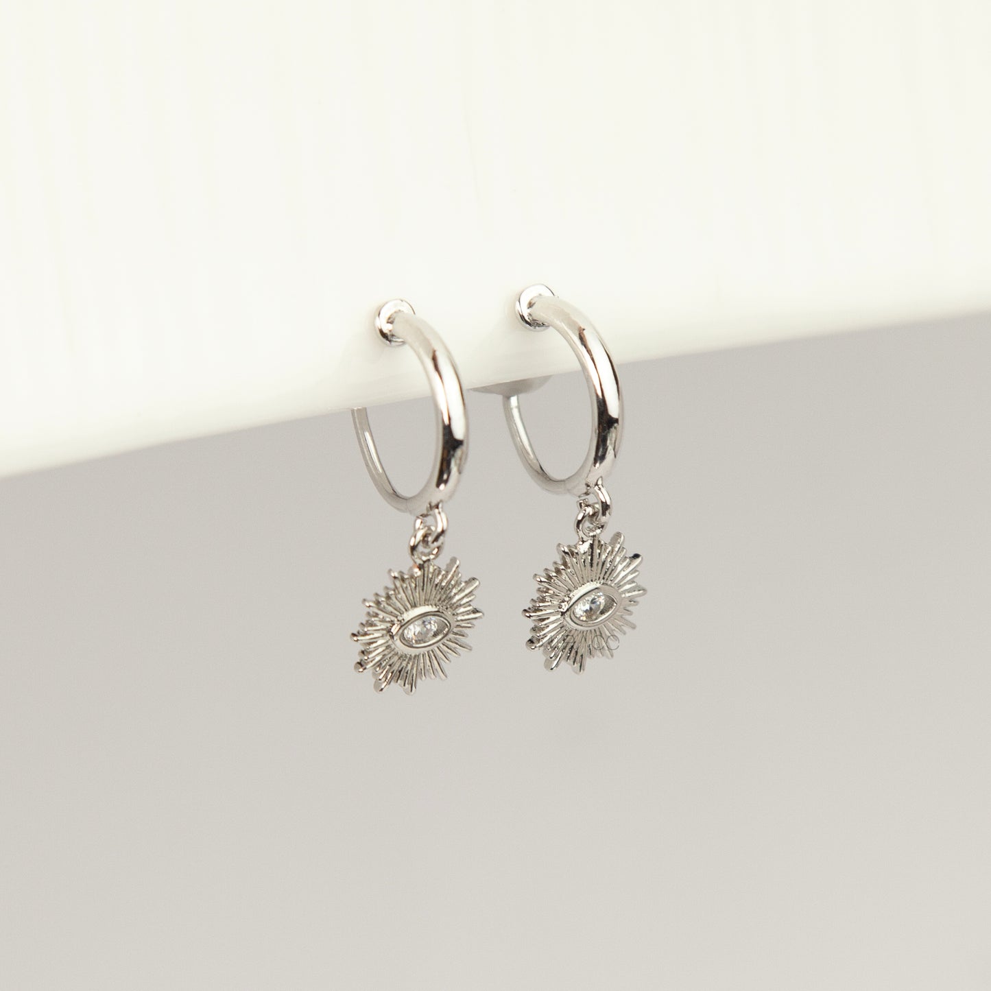 Silver coil-back clip-on hoop earrings featuring a dangling sunburst charm with a central oval crystal evil eye motif 